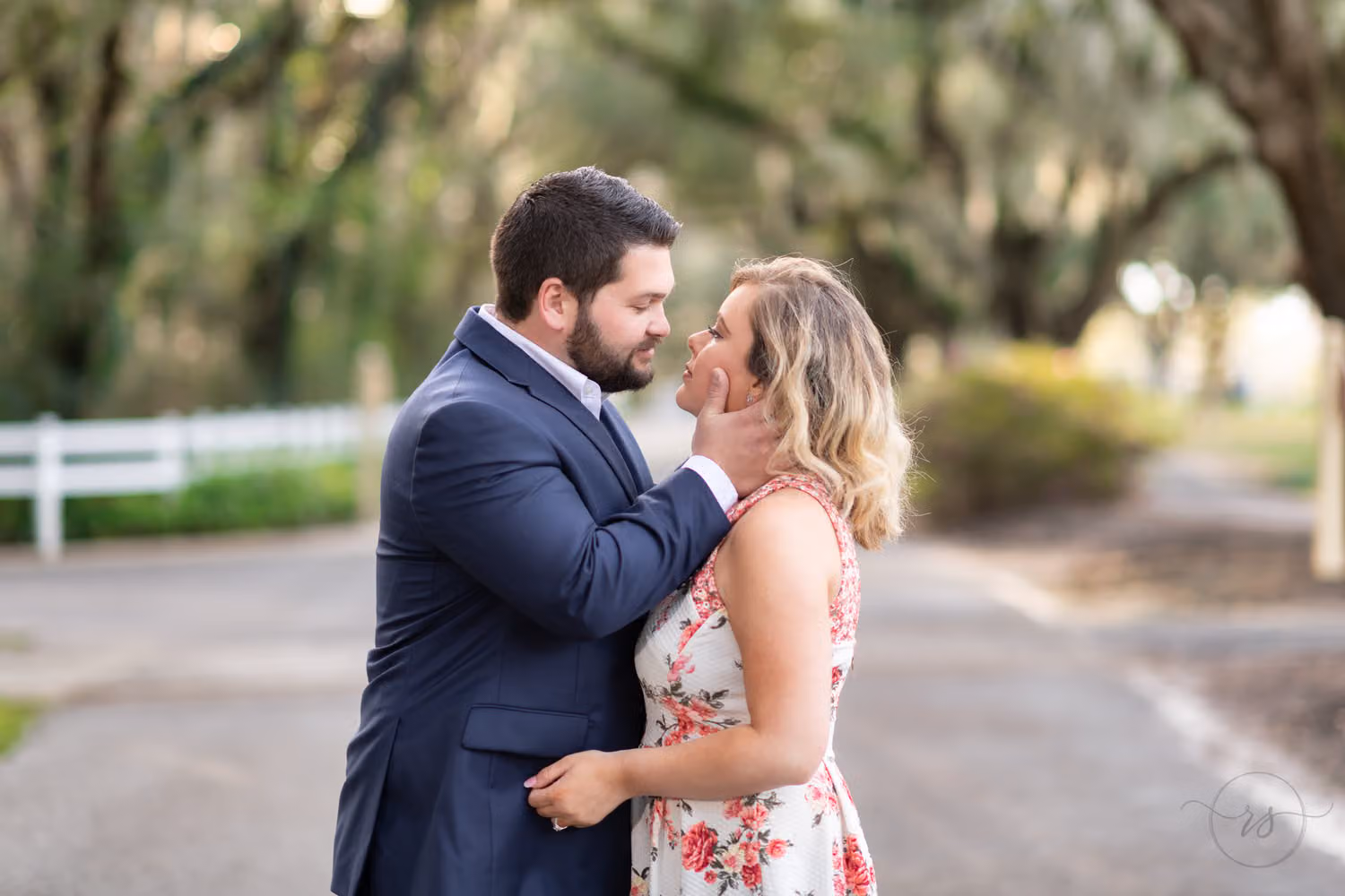 Man in a navy suit pulling his fiancée in close for a kiss beneath oak trees at Caledonia Golf & Fish Club