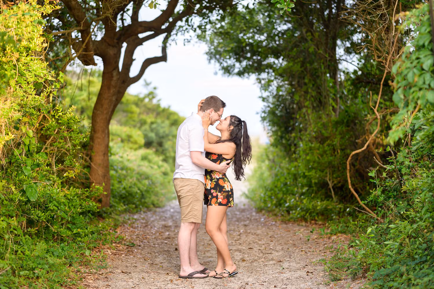 Couple kissing on a sandy walkway surrounded by green trees and bushes at Huntington Beach State Park