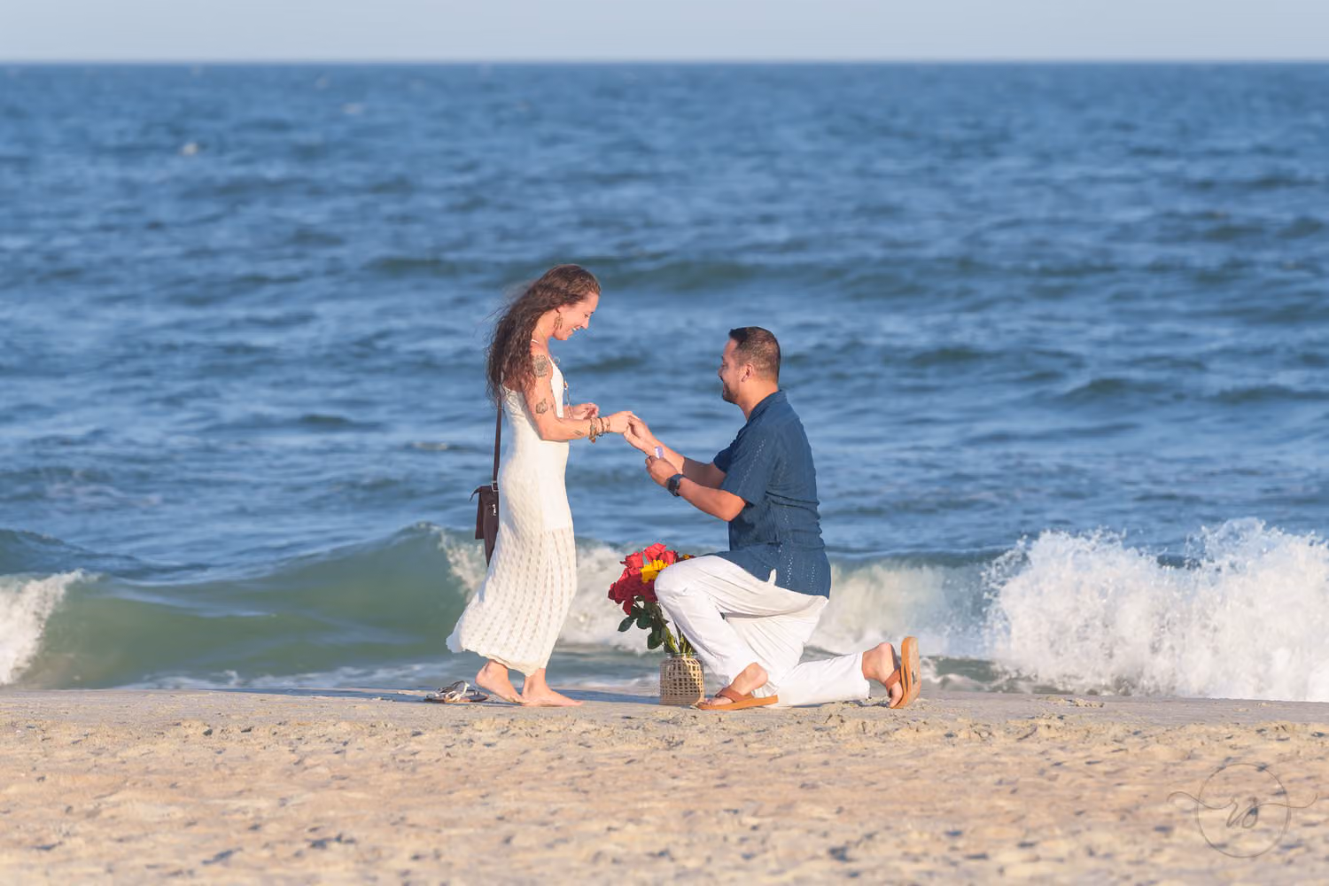 Man on one knee slipping the ring on her finger as a wave splashes behind them at Huntington Beach State Park