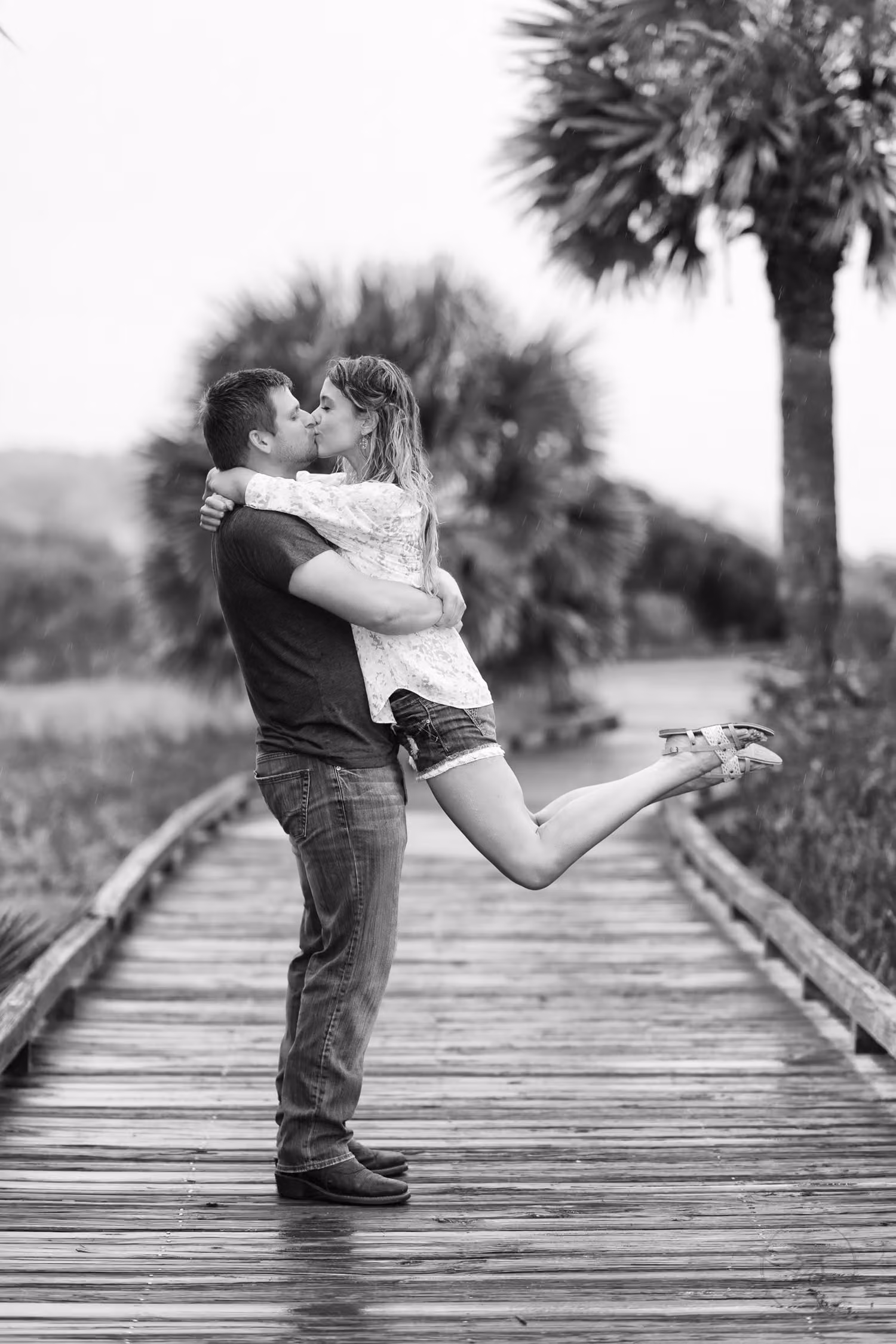 Black and white photo of a man hugging his fiancée as she kicks up a heel on a wet boardwalk at Myrtle Beach State Park
