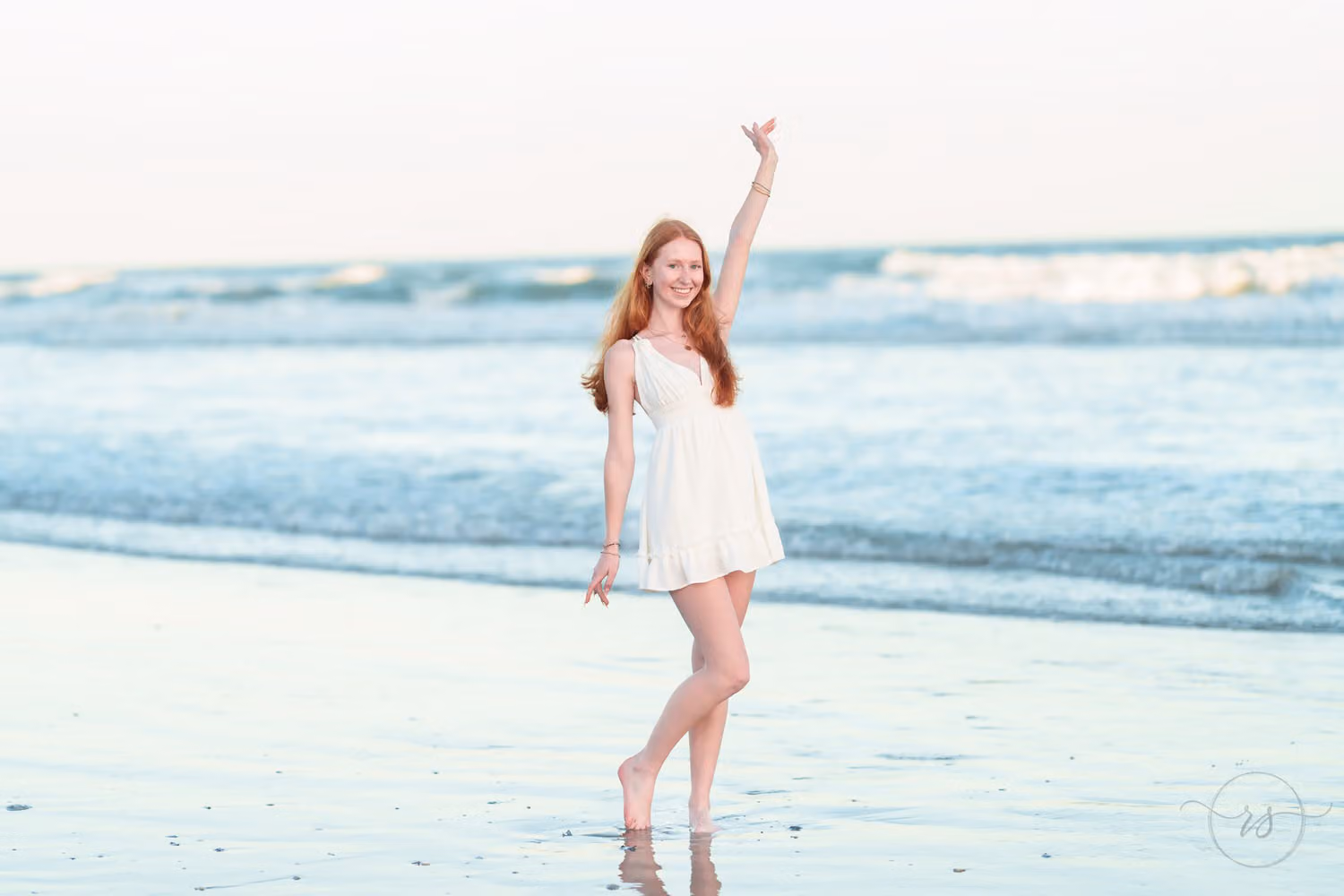 Red-haired senior girl in a white dress reaching one arm up as she stands by the waves in North Myrtle Beach