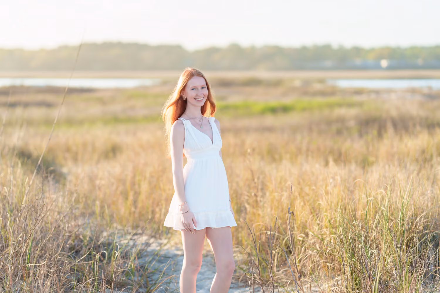 Red-haired senior girl in a white dress standing in the tall marsh grass by the water in North Myrtle Beach