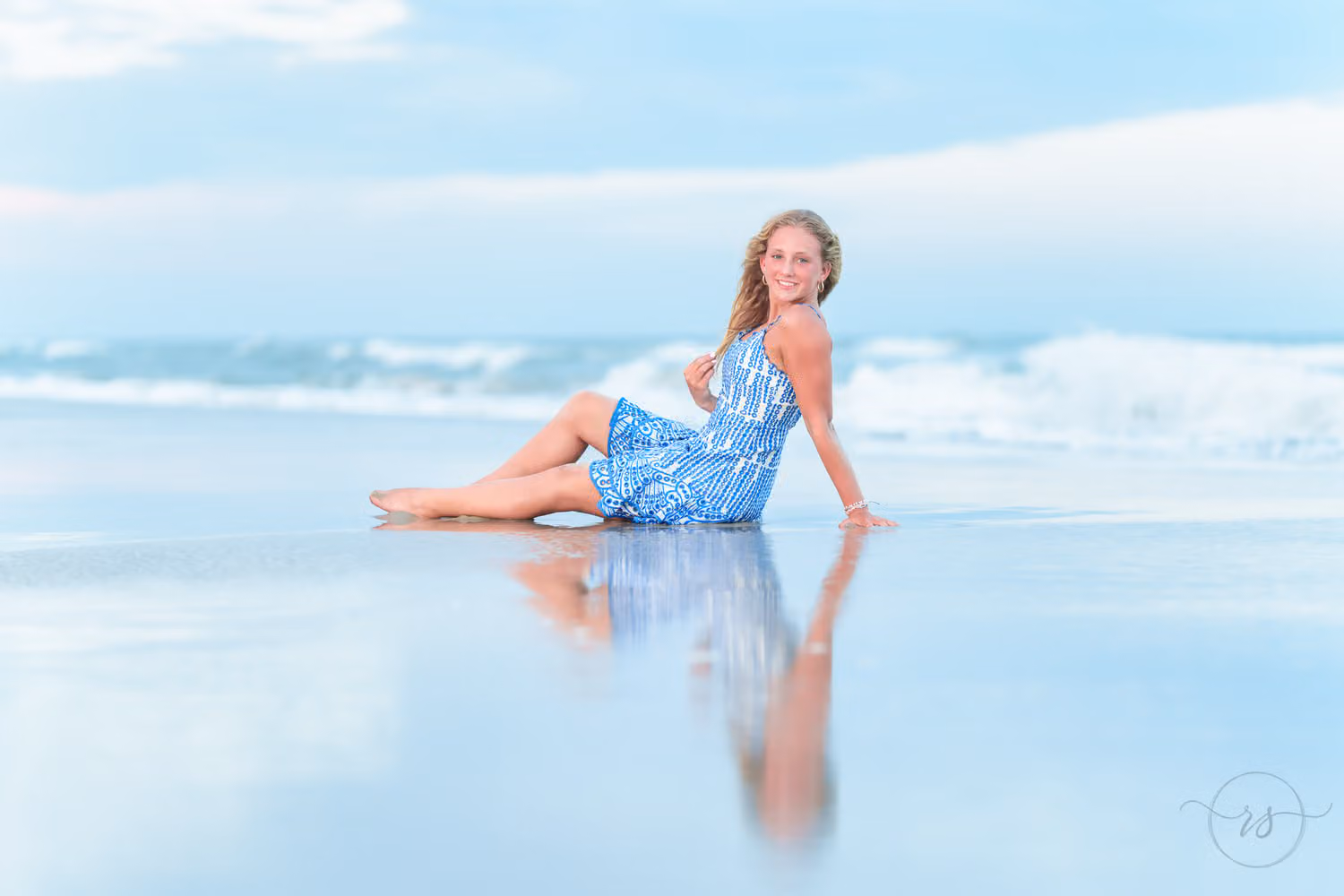 Senior girl in a blue dress reclining at the water's edge with her reflection in the wet sand at Huntington Beach State Park