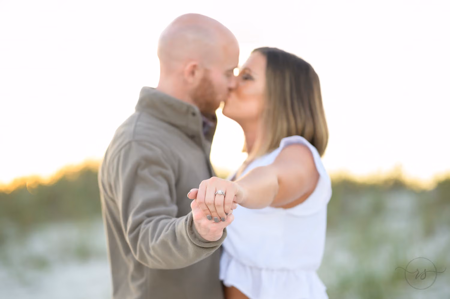Couple kissing while she holds out her hand so the engagement ring is in sharp focus at Huntington Beach State Park