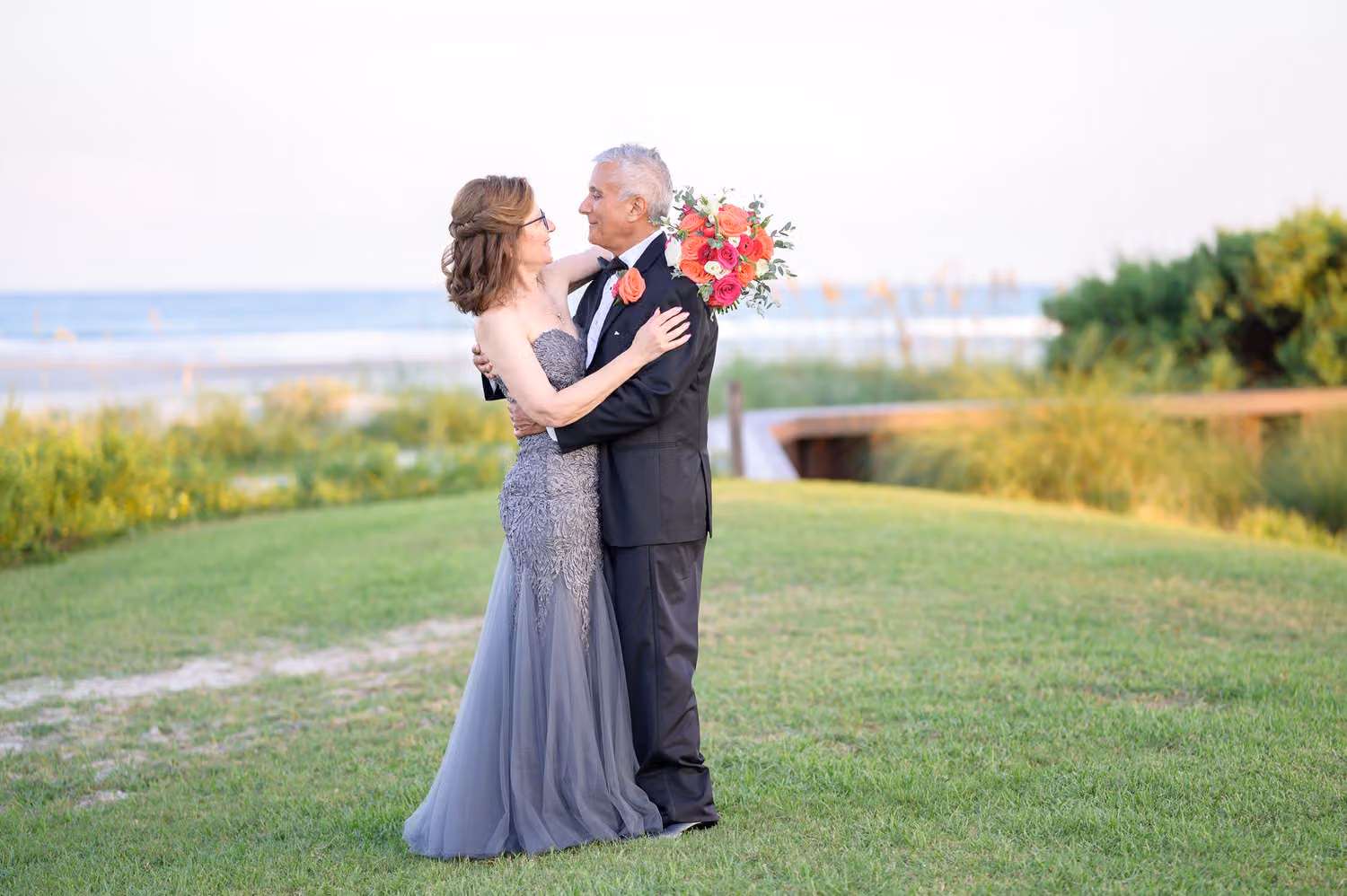 Bride in a gray gown and groom embracing on the lawn above the beach at Dunes Golf & Beach Club