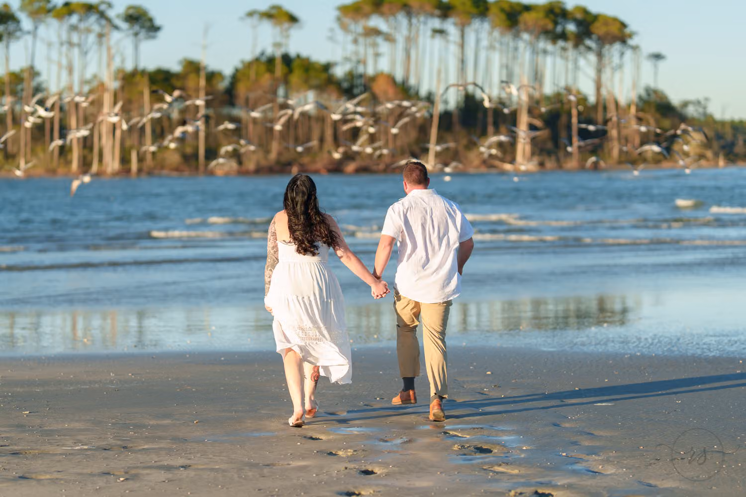Couple running hand in hand across the wet sand toward the inlet as a flock of seagulls flies up