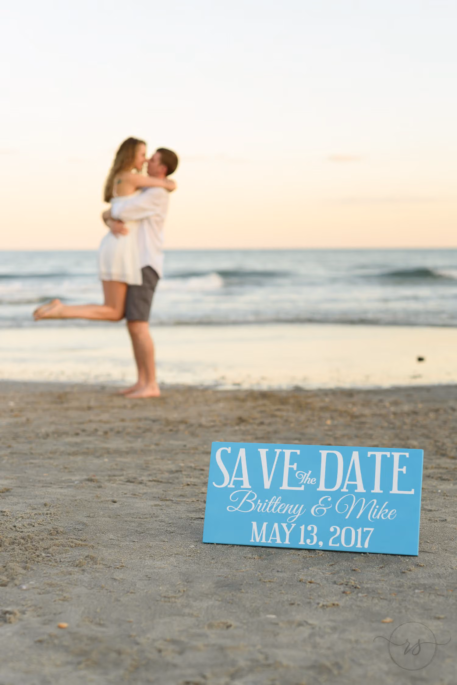 Couple hugging by the ocean with a blue save the date sign propped in the sand at Myrtle Beach State Park