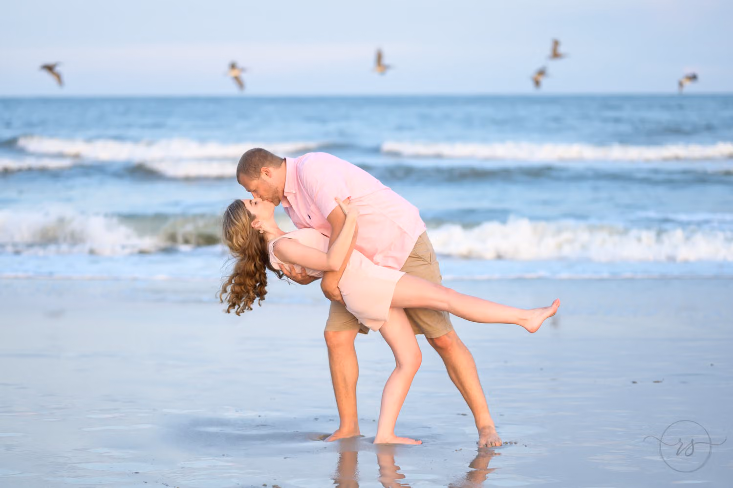 Man dipping his fiancée for a kiss in the shallow water as seagulls fly overhead at Huntington Beach State Park