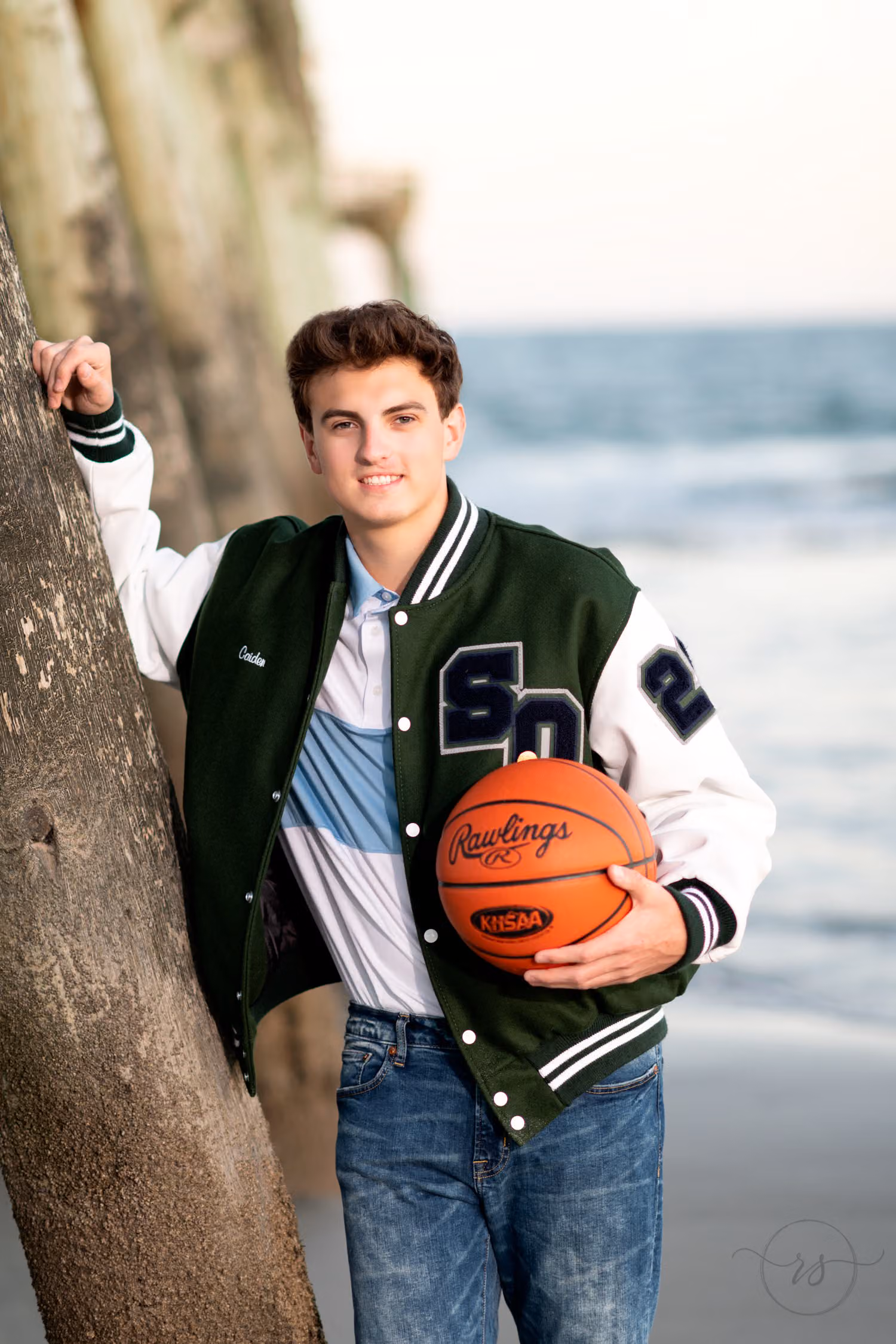 Senior in a green letterman jacket holding a basketball and leaning on a pier piling at Myrtle Beach State Park