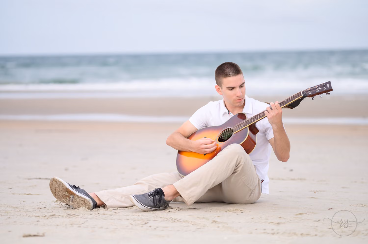 Senior guy sitting in the sand playing an acoustic guitar with the ocean behind him at Huntington Beach State Park