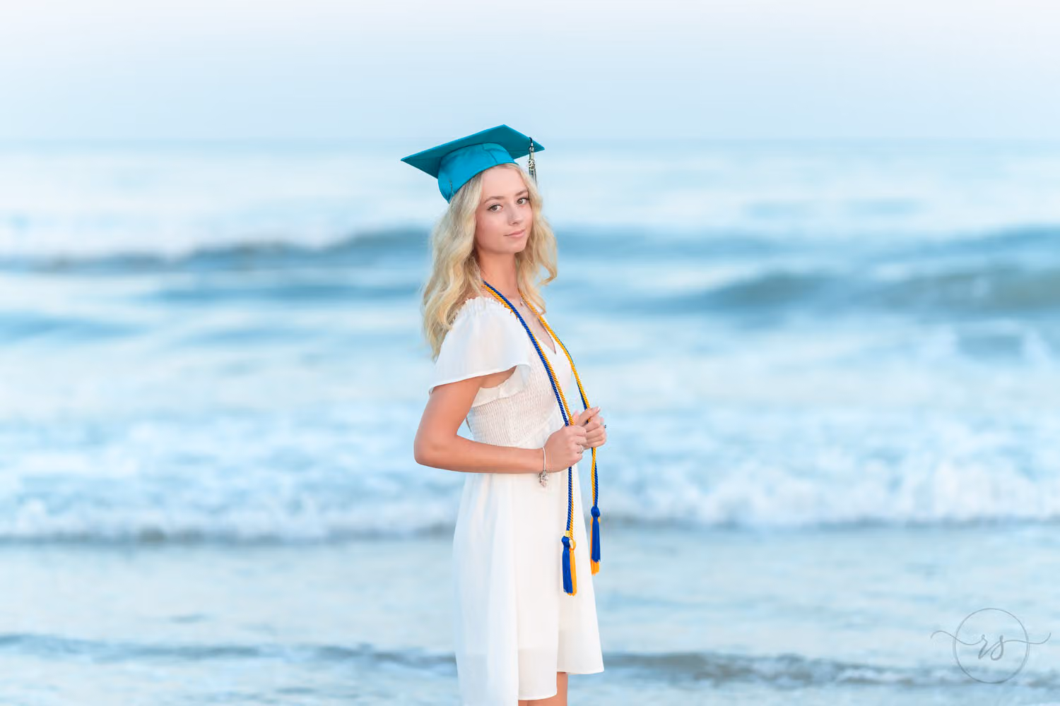 Senior in a white dress, teal cap and graduation cords looking off to the side by the ocean at Huntington Beach State Park