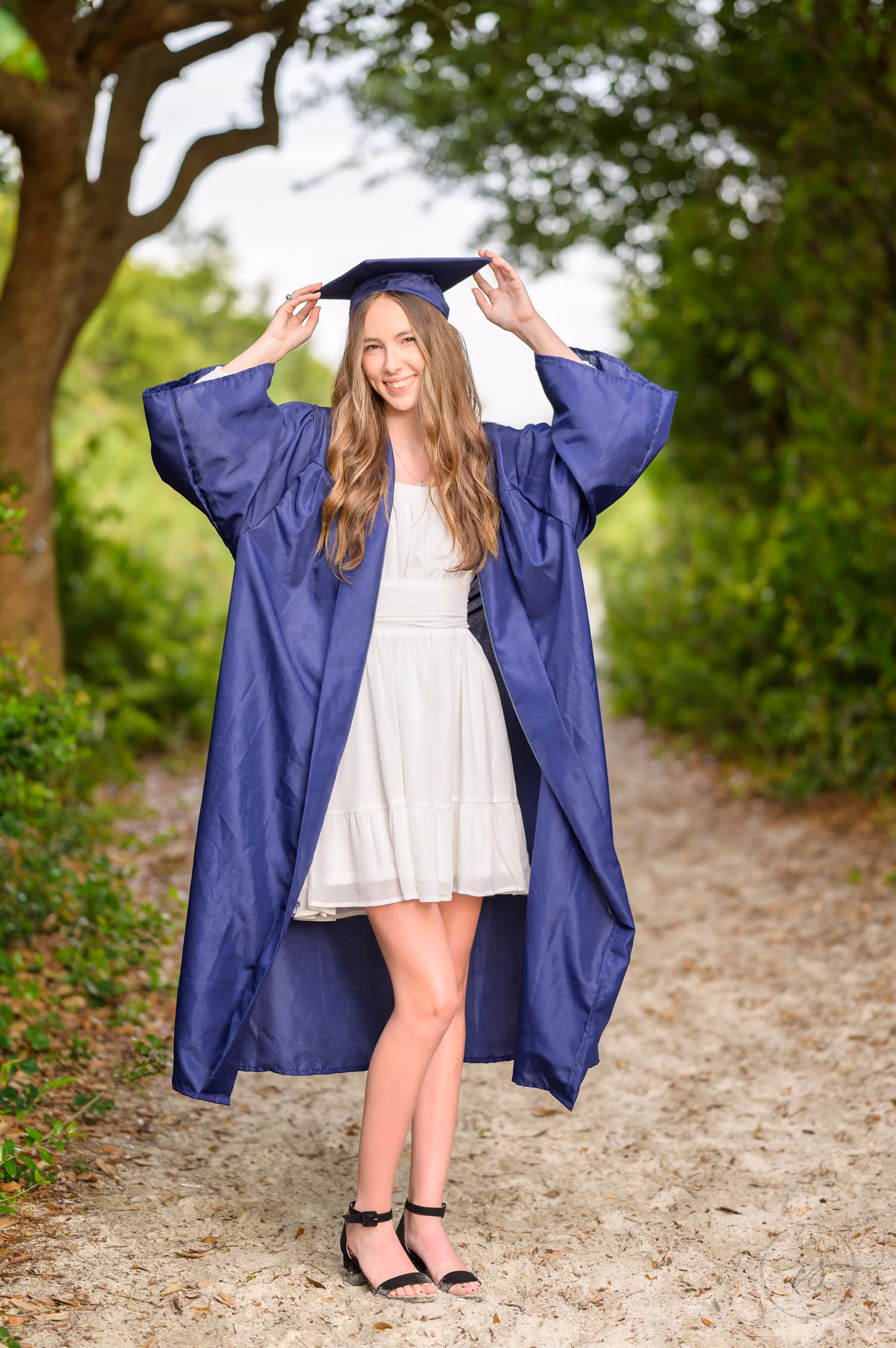 Senior in a navy cap and gown holding her cap on a sandy path under the trees at Huntington Beach State Park