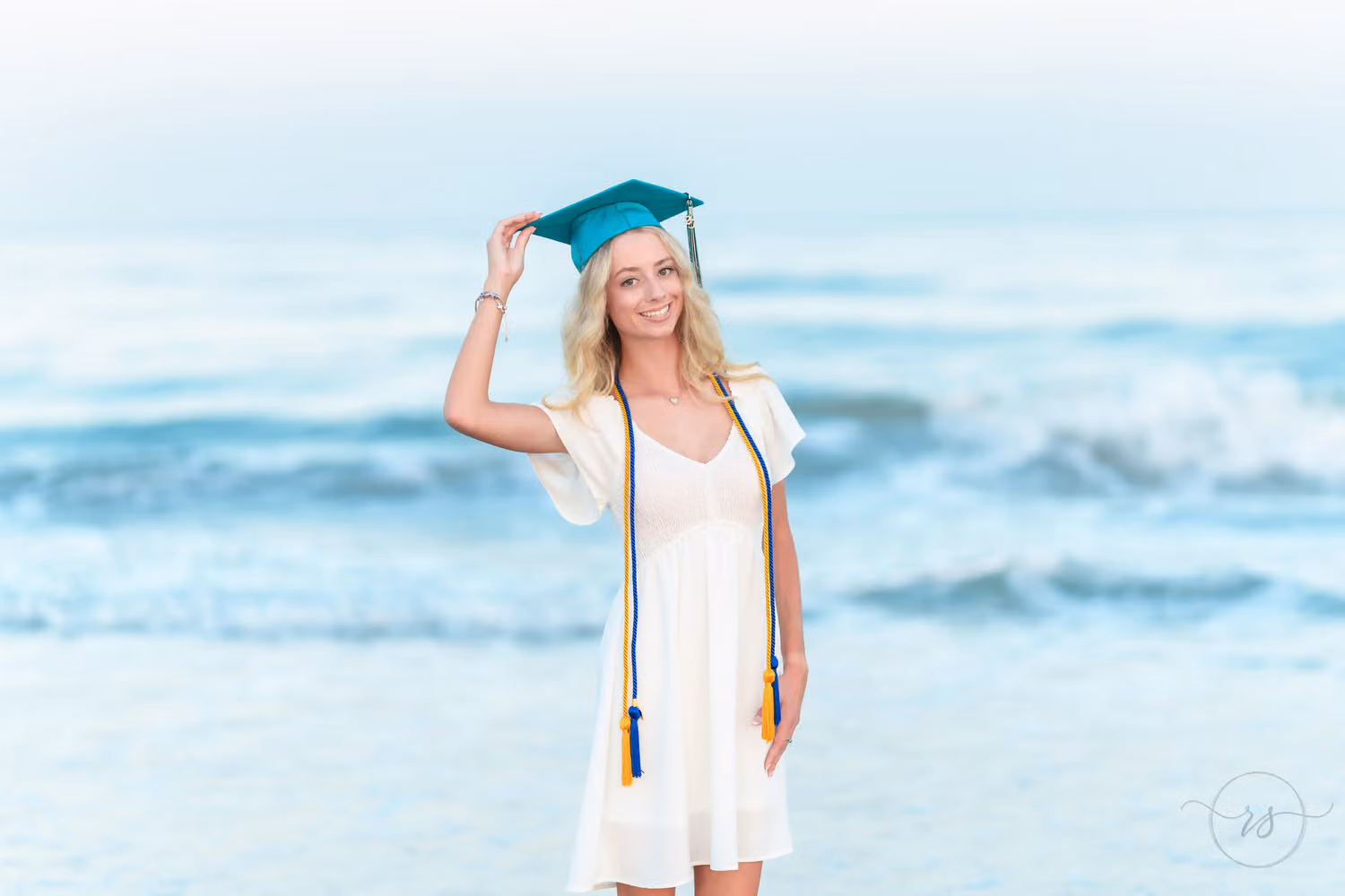 Senior in a white dress and graduation cords holding her teal cap by the waves at Huntington Beach State Park