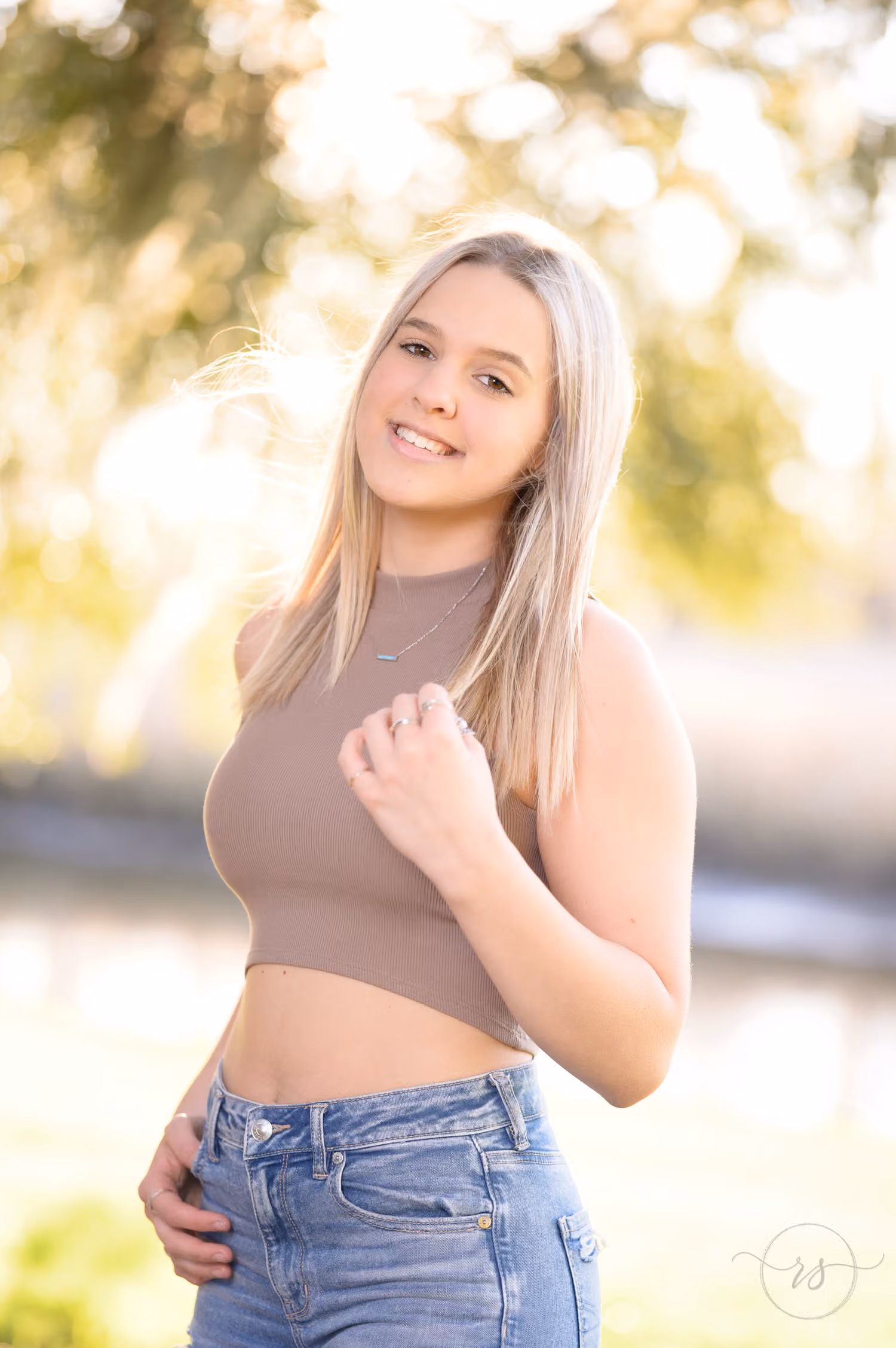 Blonde senior girl in jeans and a crop top smiling with warm sunset light glowing behind her at Caledonia Golf & Fish Club