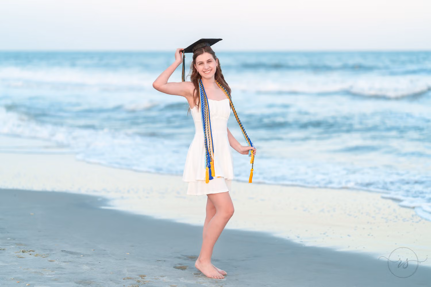 Senior girl in a white dress and gold cords holding her graduation cap on by the ocean at Huntington Beach State Park