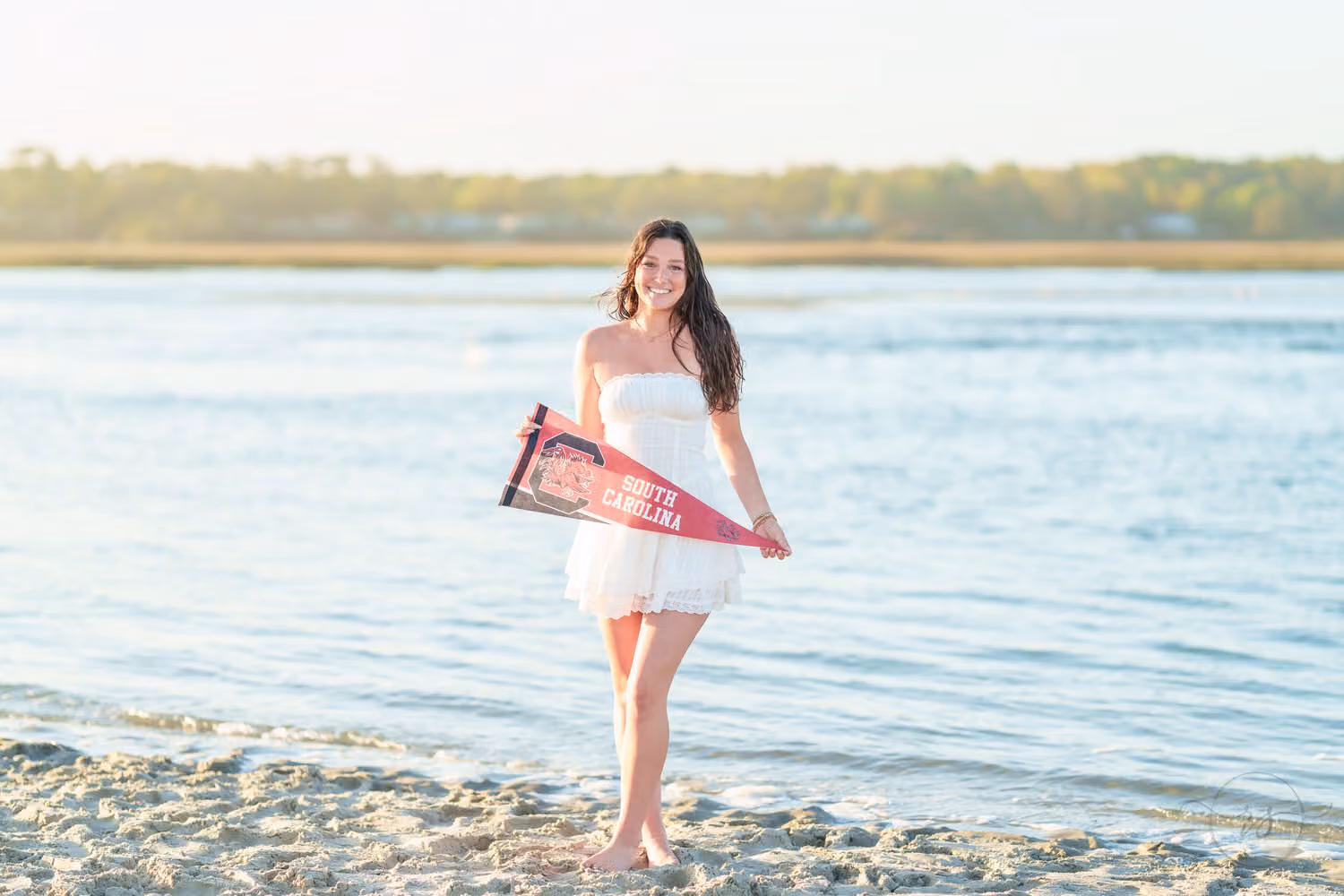 Senior girl in a white dress holding a South Carolina pennant by the water in North Myrtle Beach