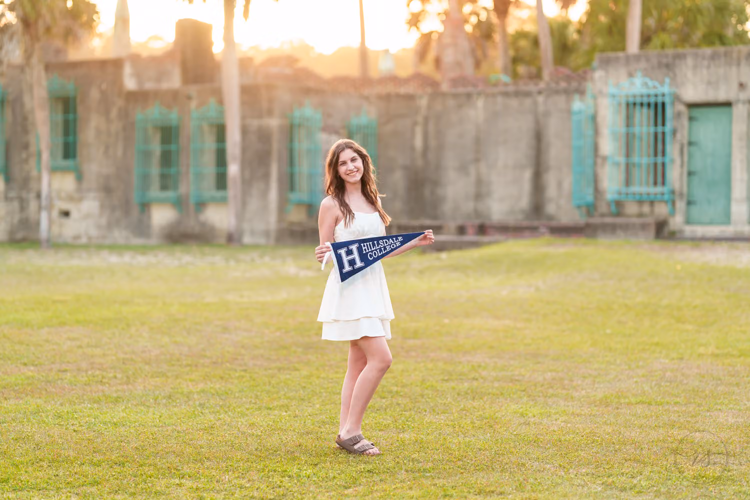 Senior girl in a white dress holding a Hillsdale College pennant on the lawn by Atalaya at Huntington Beach State Park
