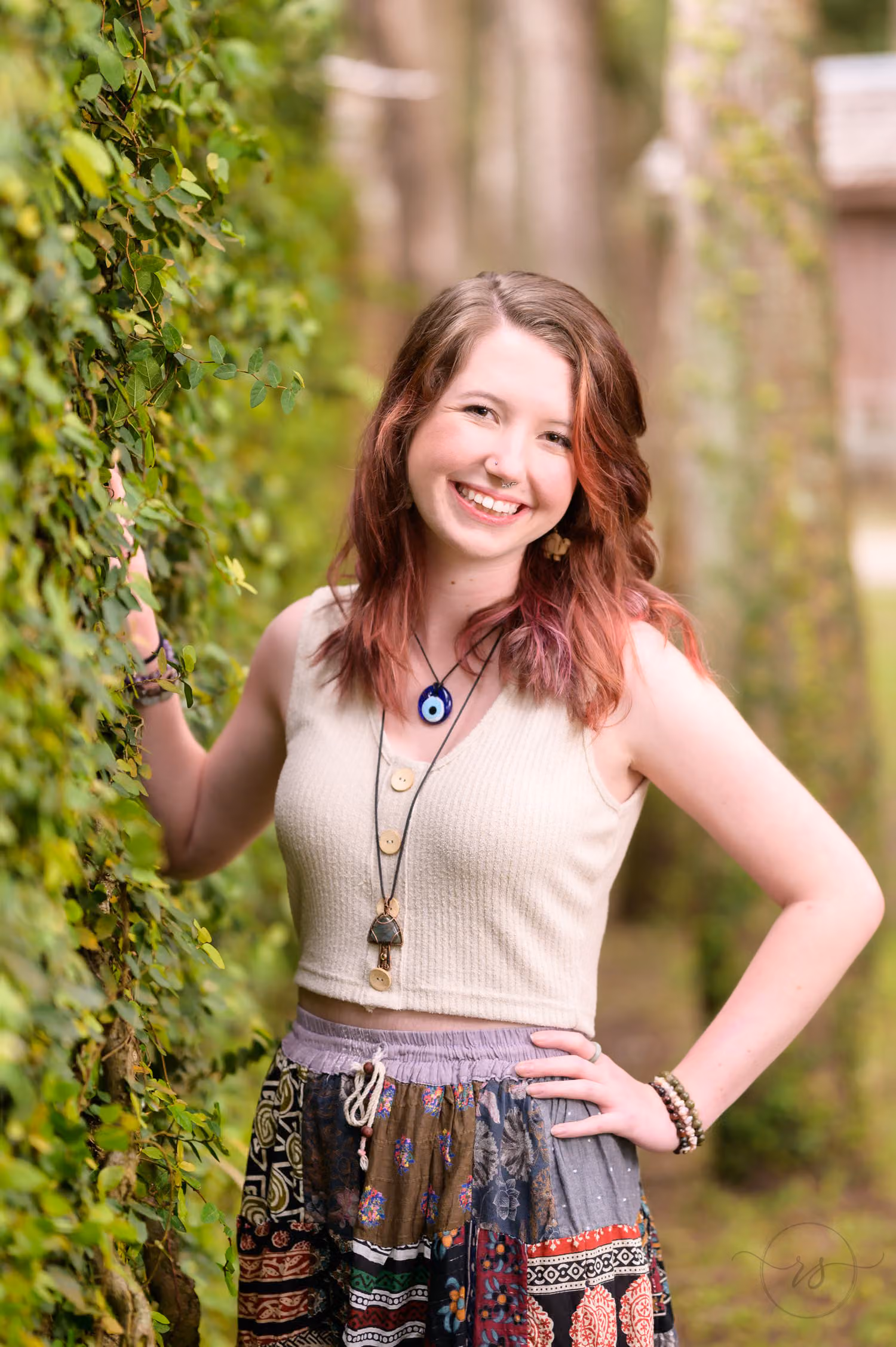 Red-haired senior girl in a patterned bohemian skirt smiling as she leans on the ivy-covered wall at Huntington Beach State Park