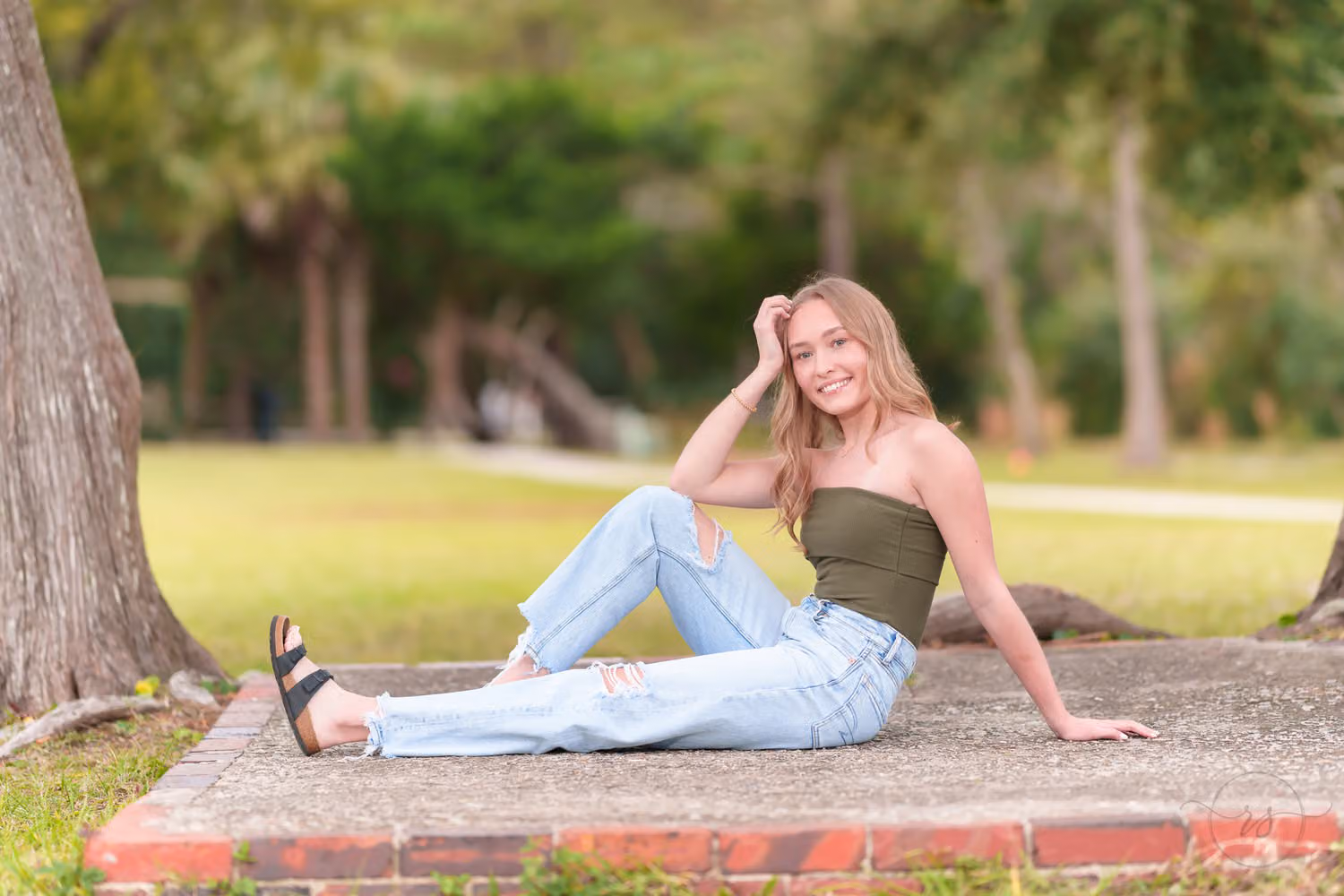 Senior girl in ripped jeans and an olive top reclining on a stone slab under the trees at Huntington Beach State Park