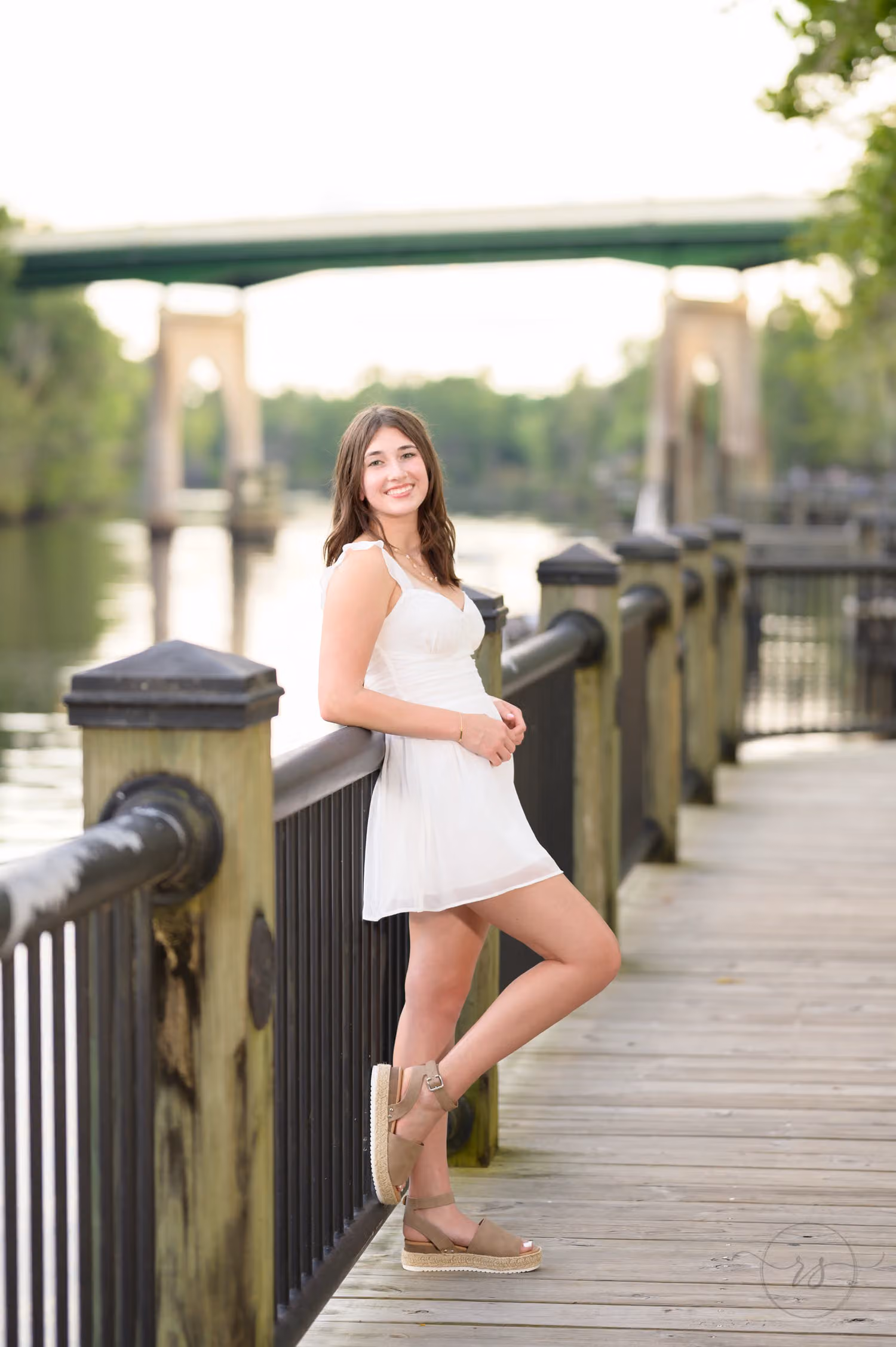 Senior girl in a white dress leaning on the boardwalk railing with the river bridge behind her at the Conway River Walk