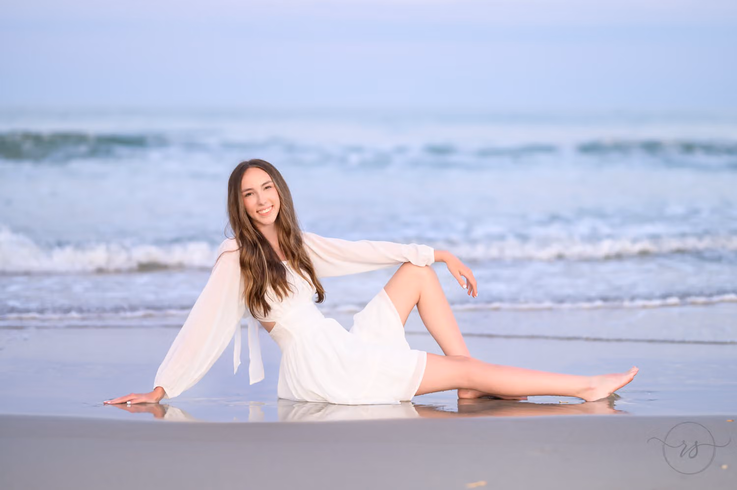Senior in a white dress reclining on the wet sand with waves rolling in behind her at Huntington Beach State Park