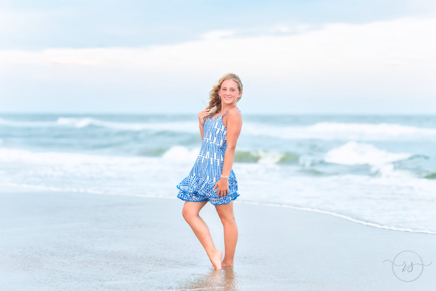 Senior girl in a blue patterned sundress standing in the shallow water by the waves at Huntington Beach State Park