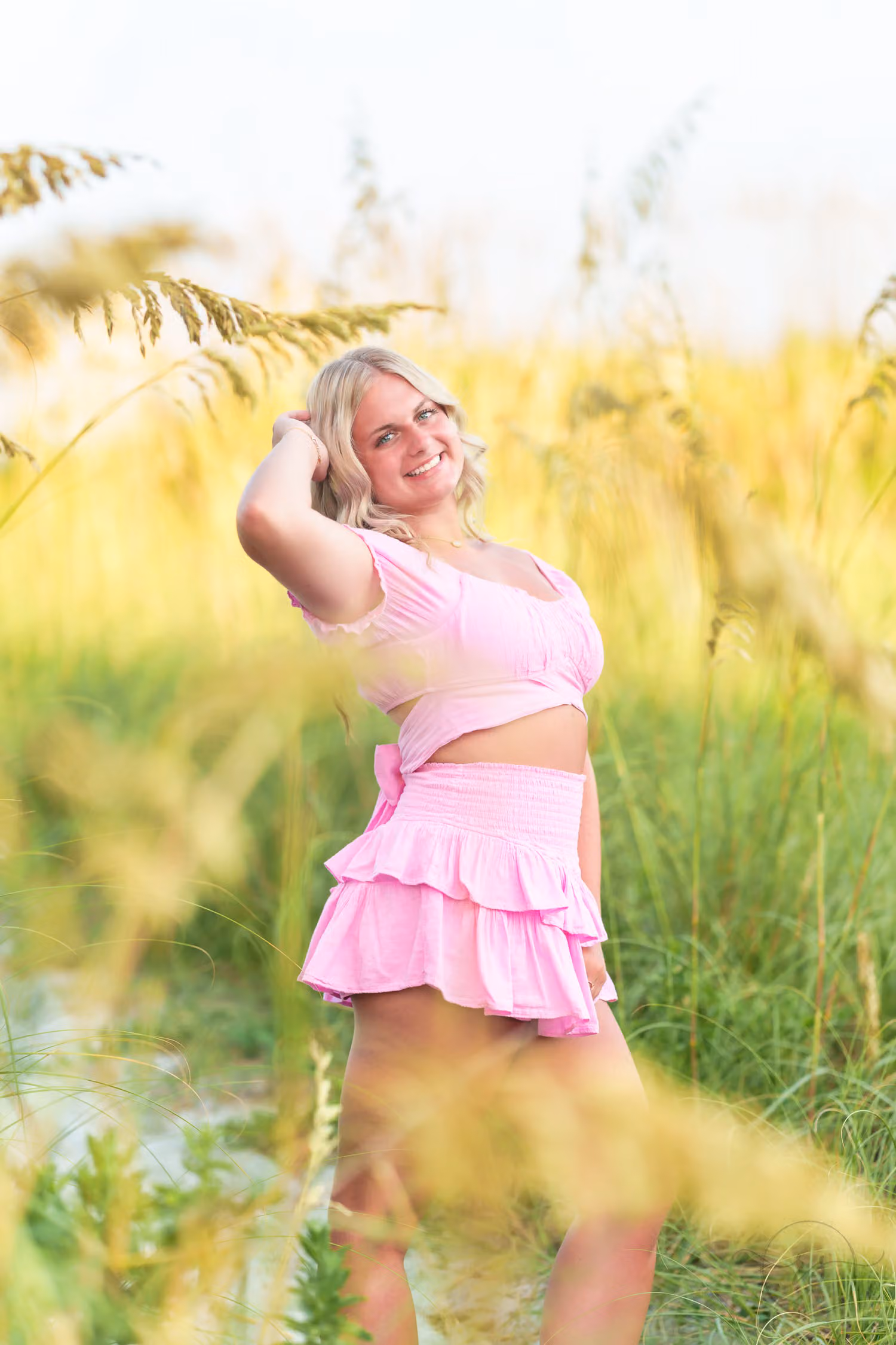 Senior girl in a pink ruffled skirt and crop top looking back over her shoulder through the sea oats in North Myrtle Beach