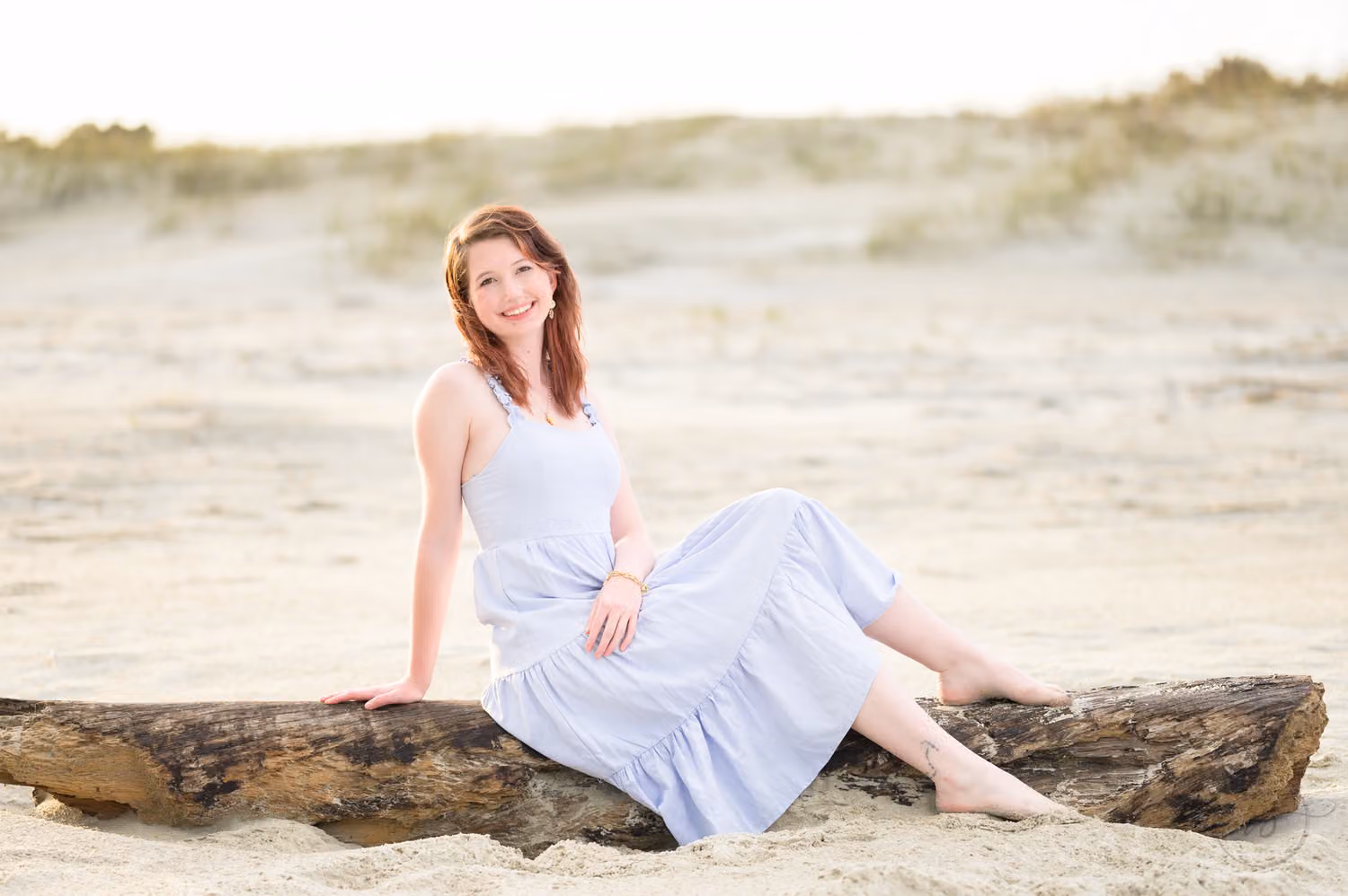 Senior girl in a light blue dress sitting on a weathered driftwood log in the sand by the dunes in Pawleys Island