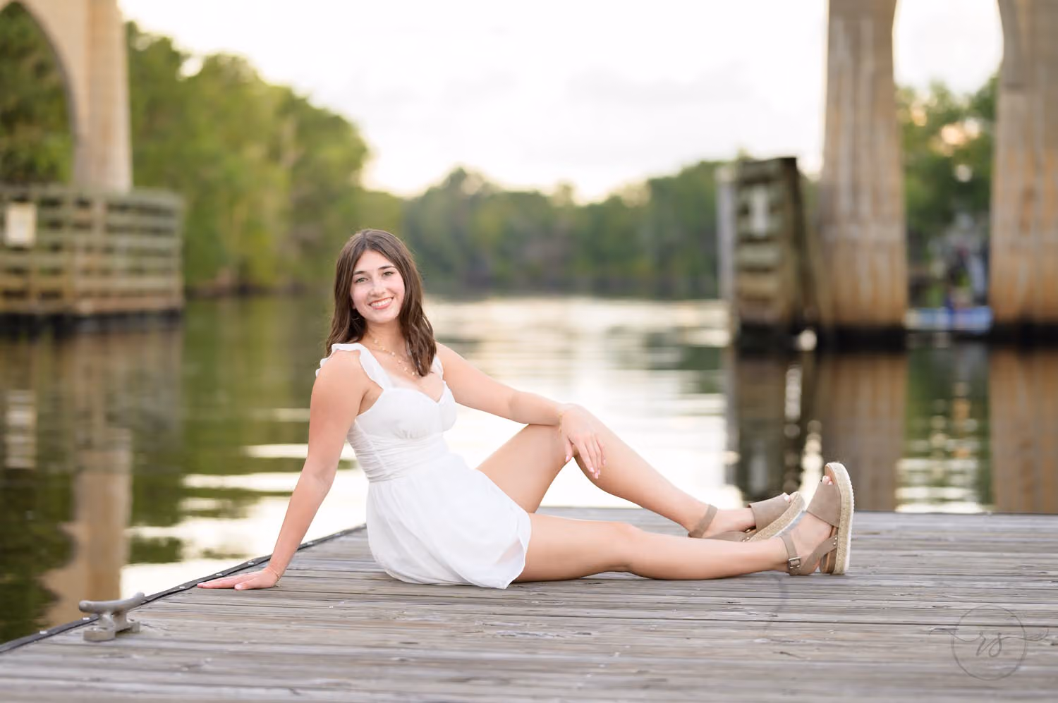 Senior girl in a white dress sitting on the wooden dock with the river behind her at the Conway River Walk