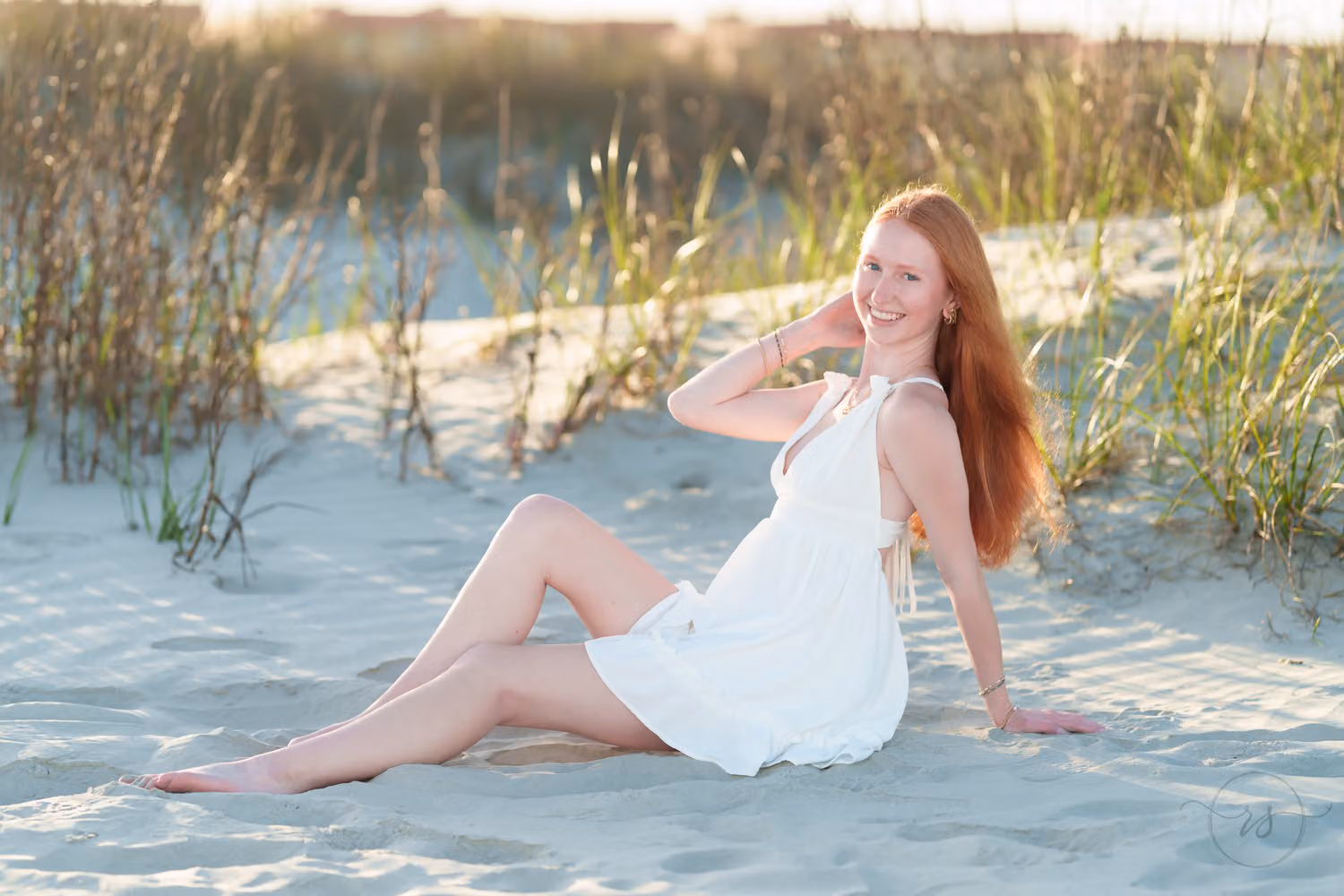 Red-haired senior girl in a white dress sitting in the sand in front of the sea oats in golden light in North Myrtle Beach