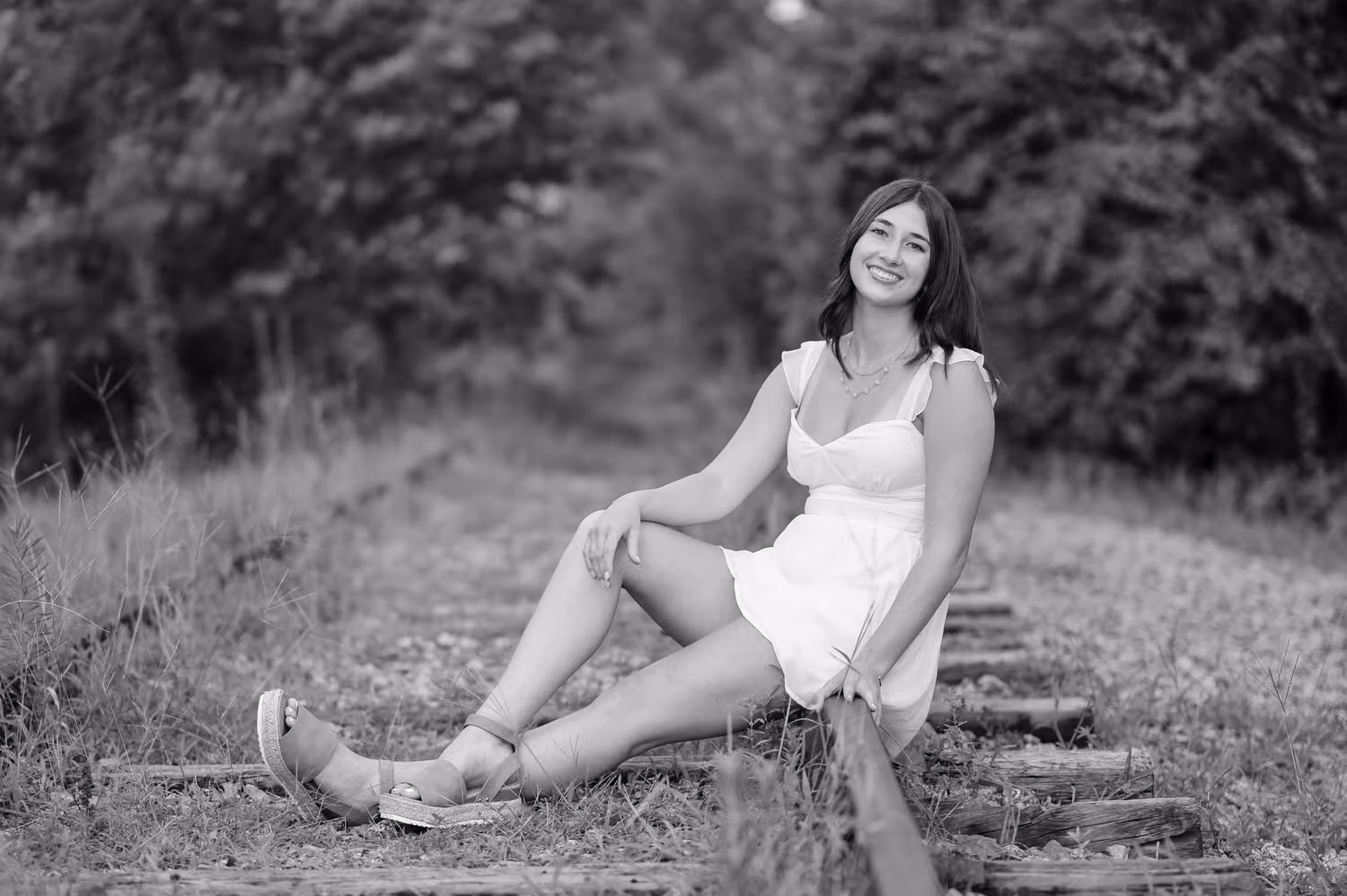 Black and white portrait of a senior girl in a white dress sitting on old train tracks by the trees at the Conway River Walk