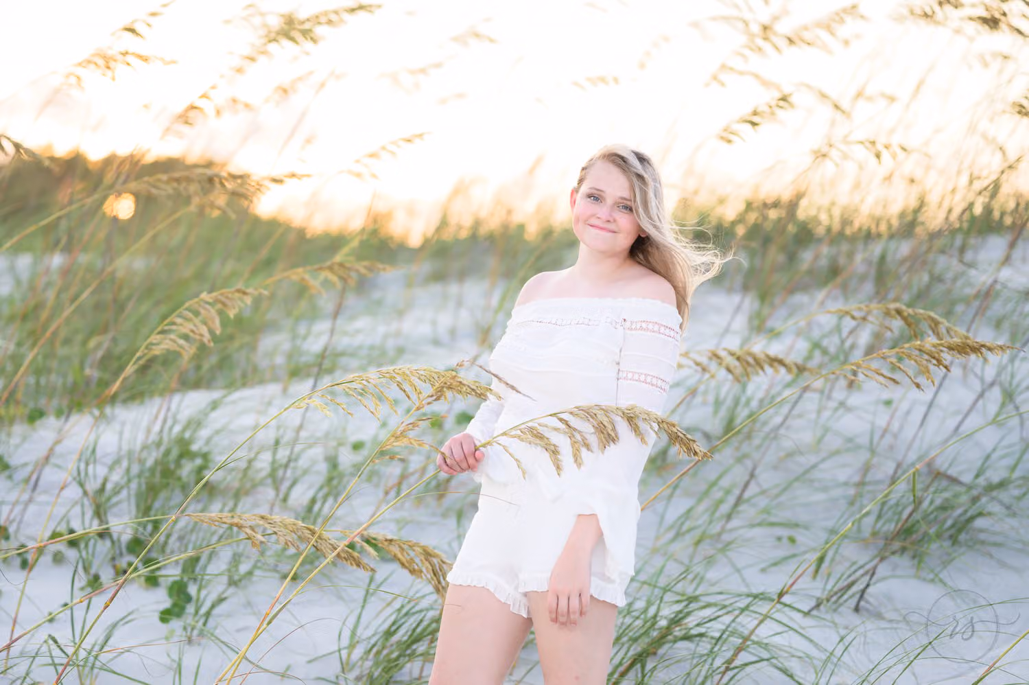 Blonde senior in an off-the-shoulder white top standing among the sea oats on the dunes at Huntington Beach State Park
