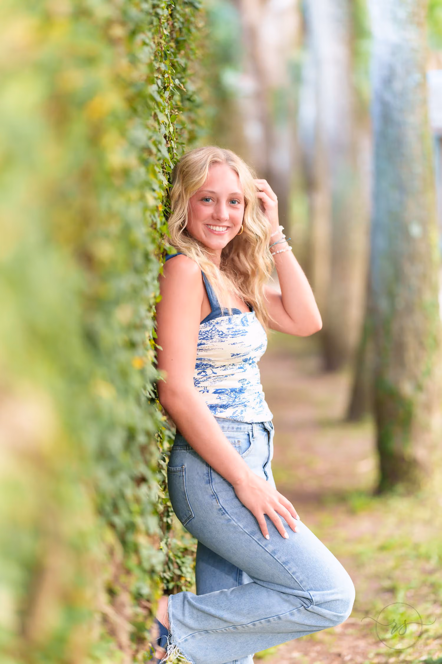 Senior girl in jeans and a blue top leaning on the ivy-covered wall with a hand in her hair at Huntington Beach State Park