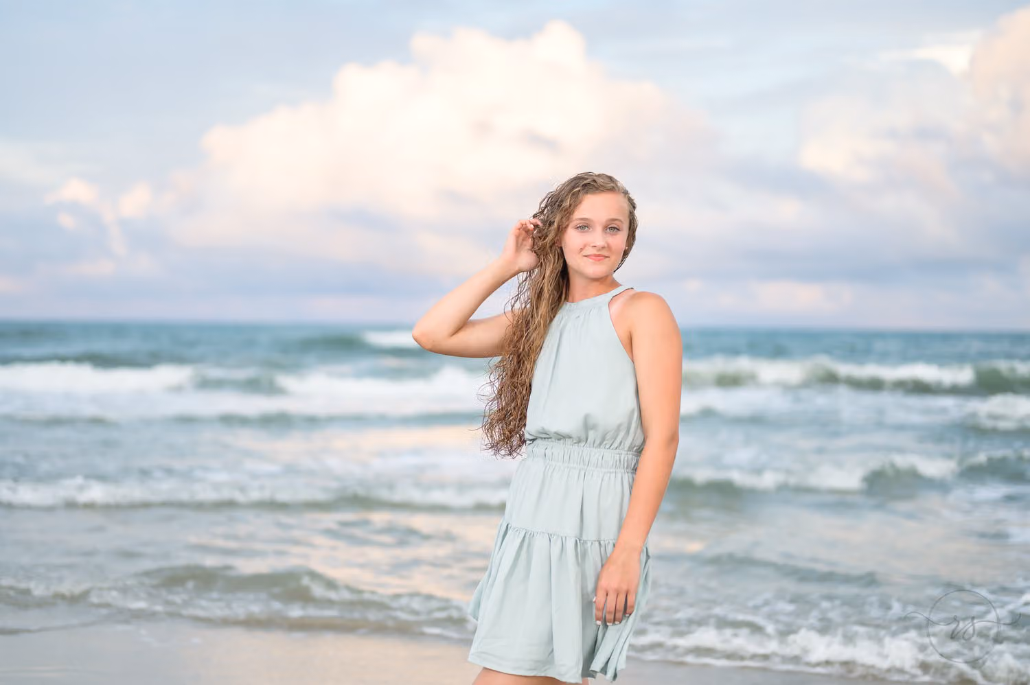 Senior girl in a mint dress touching her hair by the ocean under puffy white clouds at Huntington Beach State Park