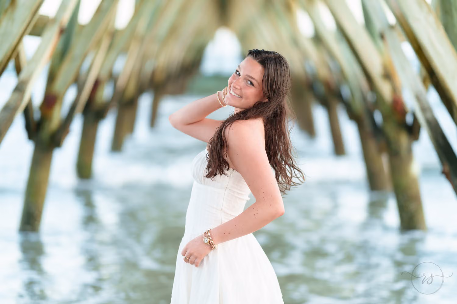 Smiling senior girl with long dark hair in a white dress posing under the pier in North Myrtle Beach