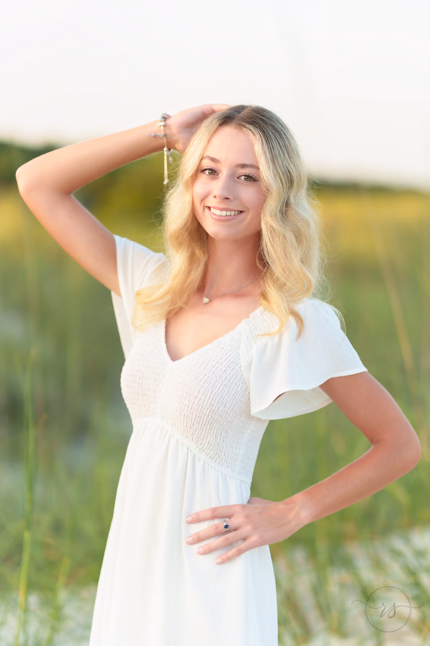 Close portrait of a smiling senior in a white dress with her hand in her hair by the dunes at Huntington Beach State Park