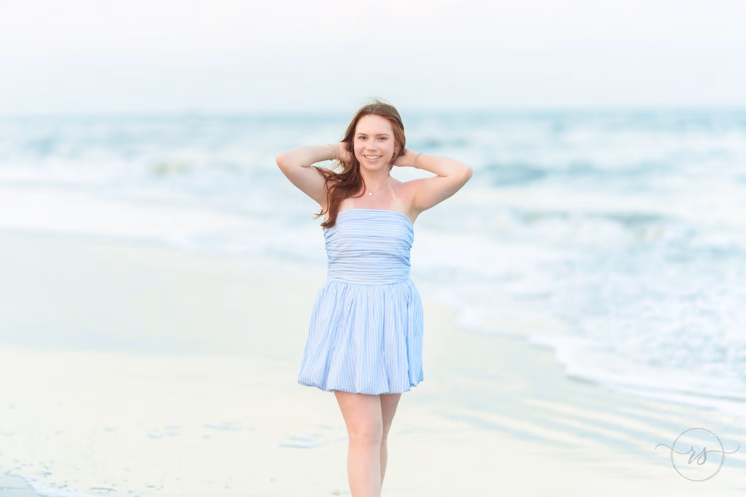 Senior girl in a light blue dress standing at the water's edge with both hands in her hair at Huntington Beach State Park