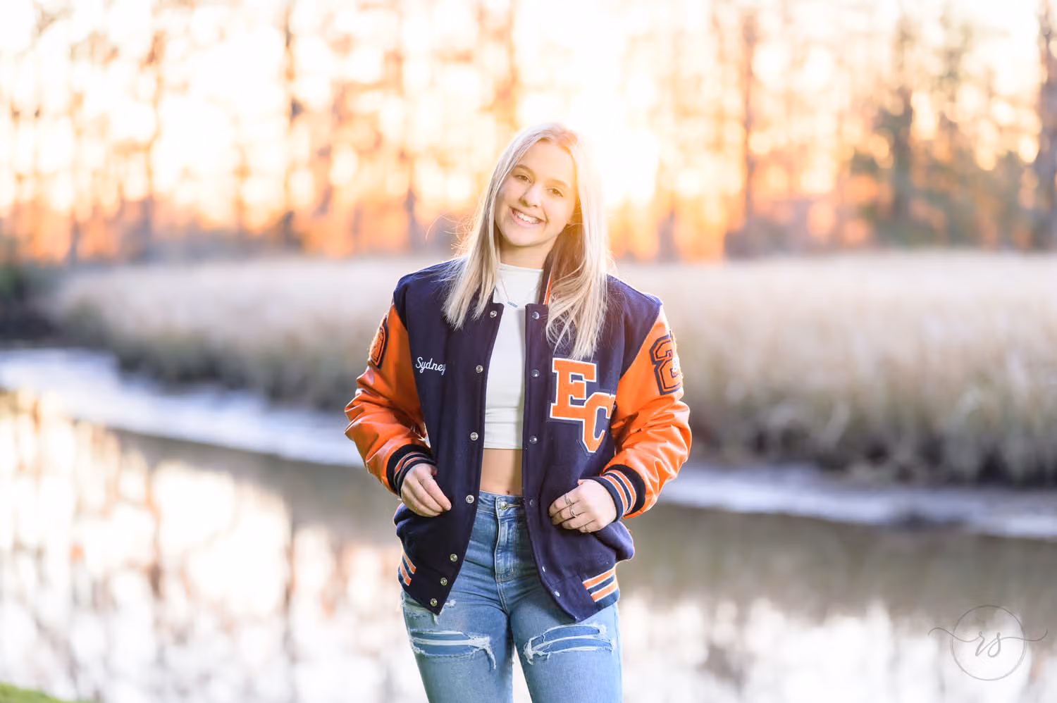 Senior girl in a letterman jacket smiling by the marsh with sunset glowing through the trees at Caledonia Golf & Fish Club