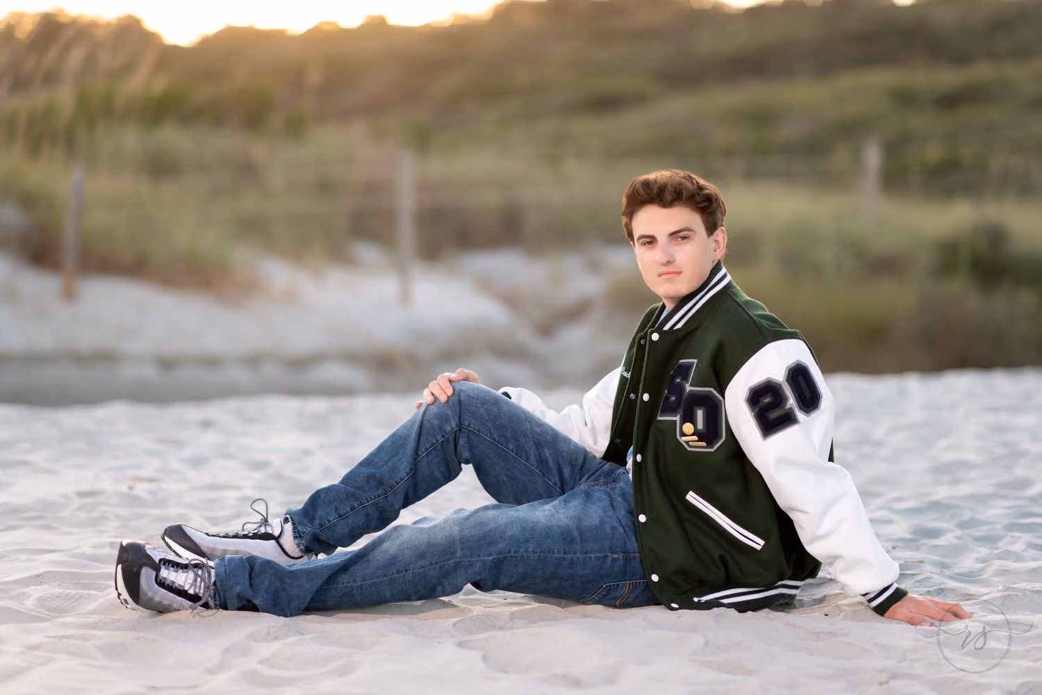 Senior in a green letterman jacket reclining on one elbow in the sand by the dunes at Myrtle Beach State Park