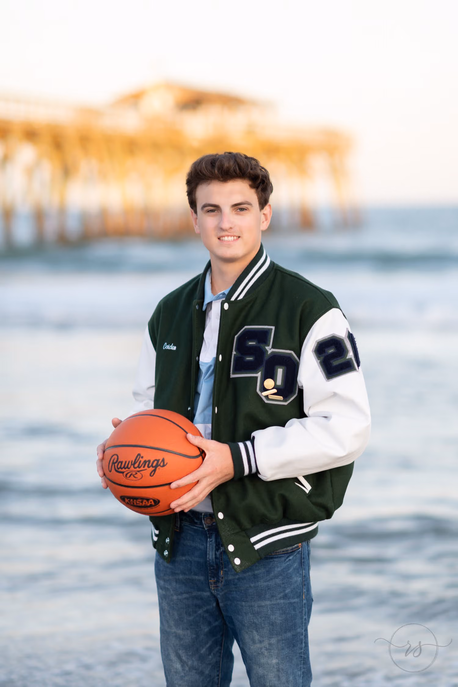 Senior in a letterman jacket holding a basketball with the pier blurred behind him at Myrtle Beach State Park
