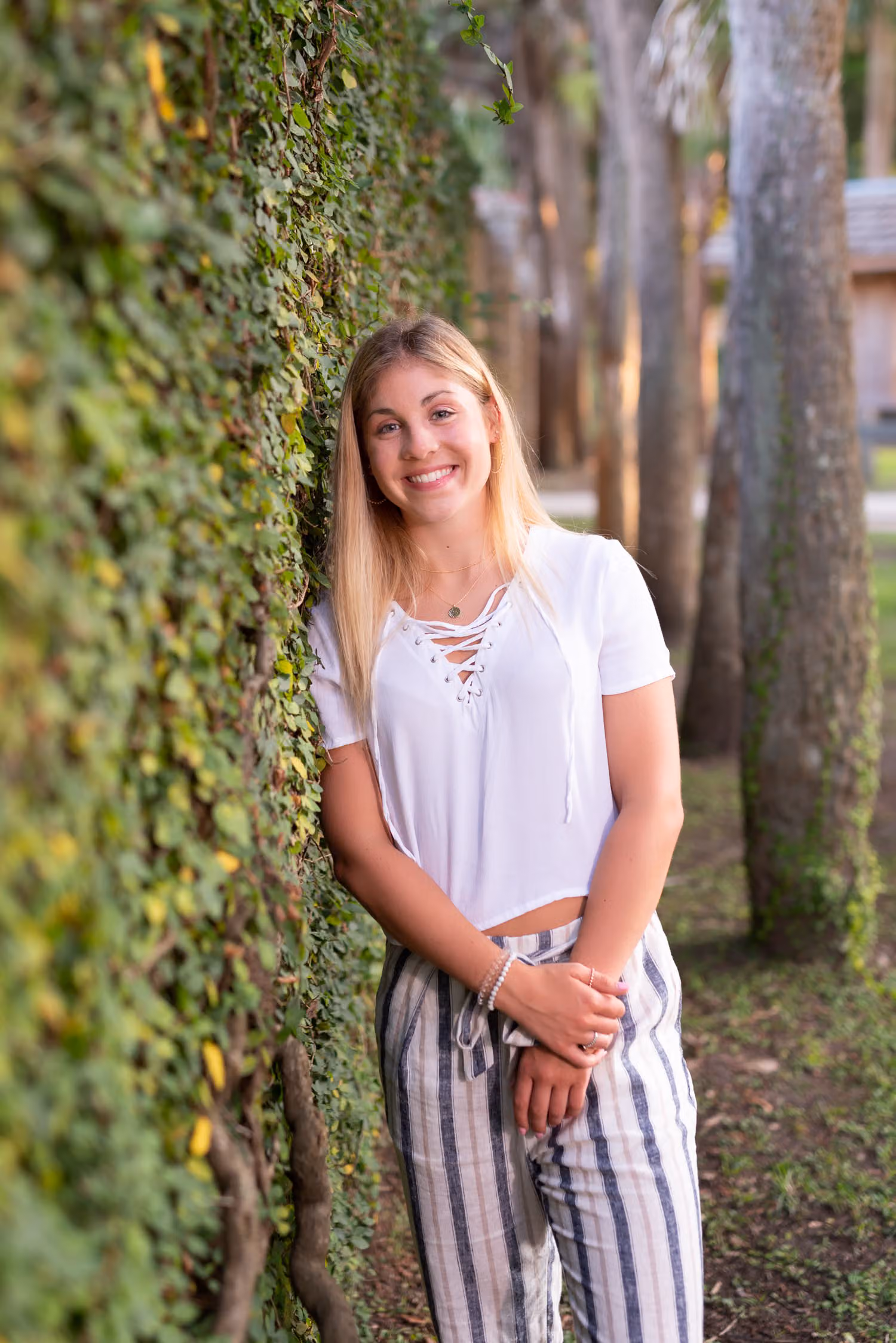 Senior in a white top and striped pants leaning against the ivy wall at Atalaya Castle