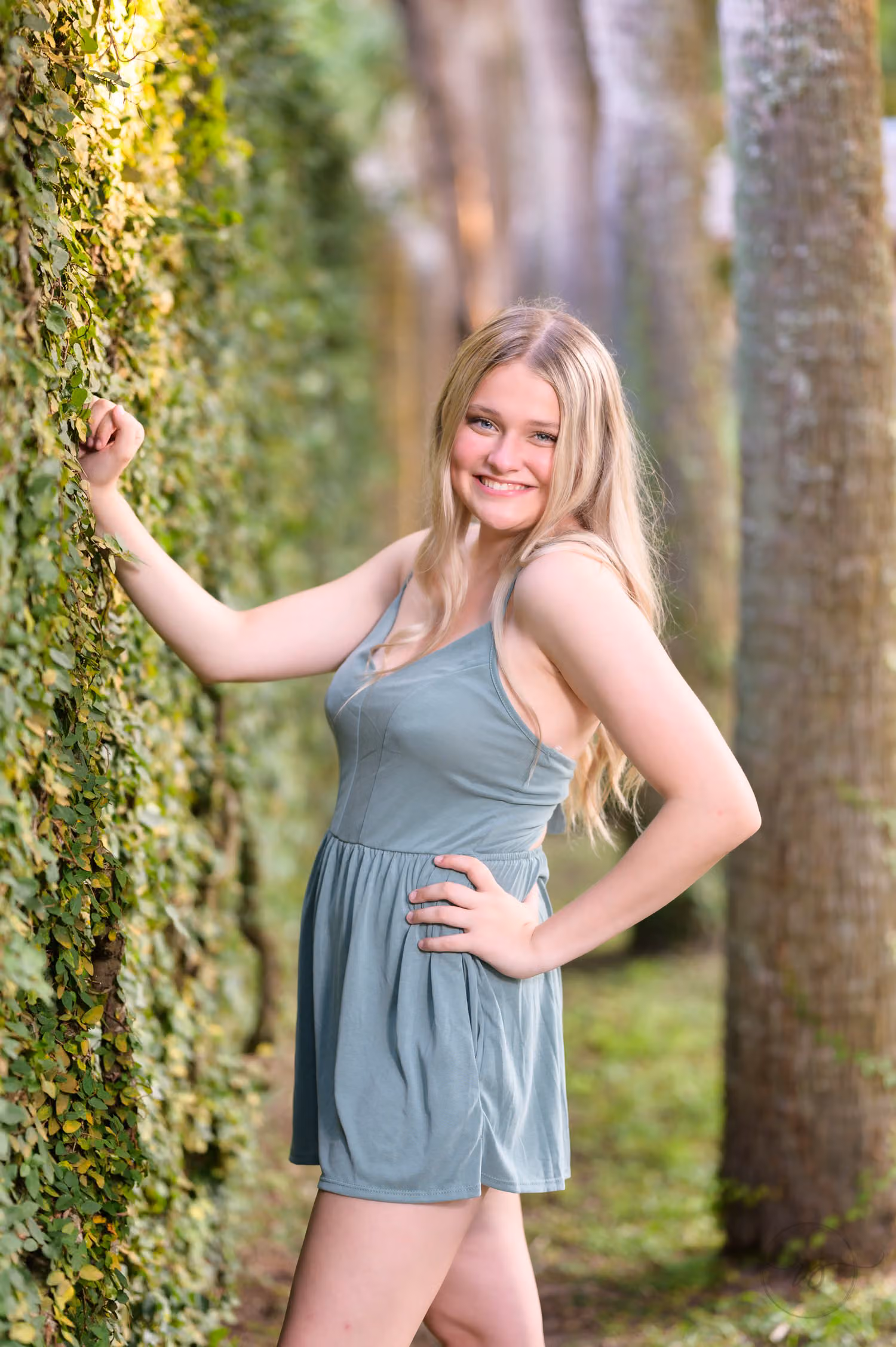 Blonde senior in a sage green romper leaning against an ivy-covered wall beside palm trunks at Huntington Beach State Park