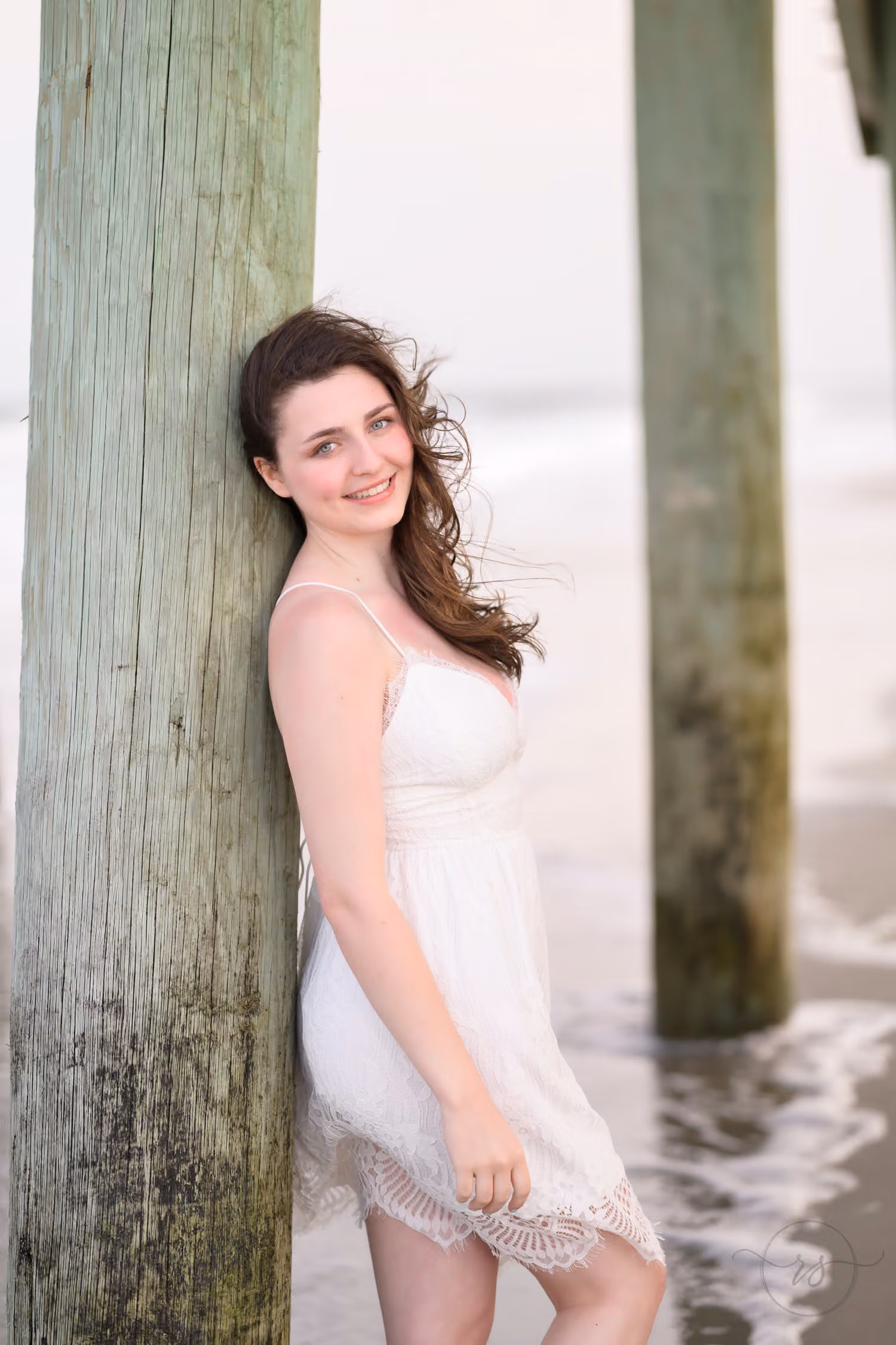 Smiling senior in a white lace dress leaning on a weathered pier piling at the water at Myrtle Beach State Park