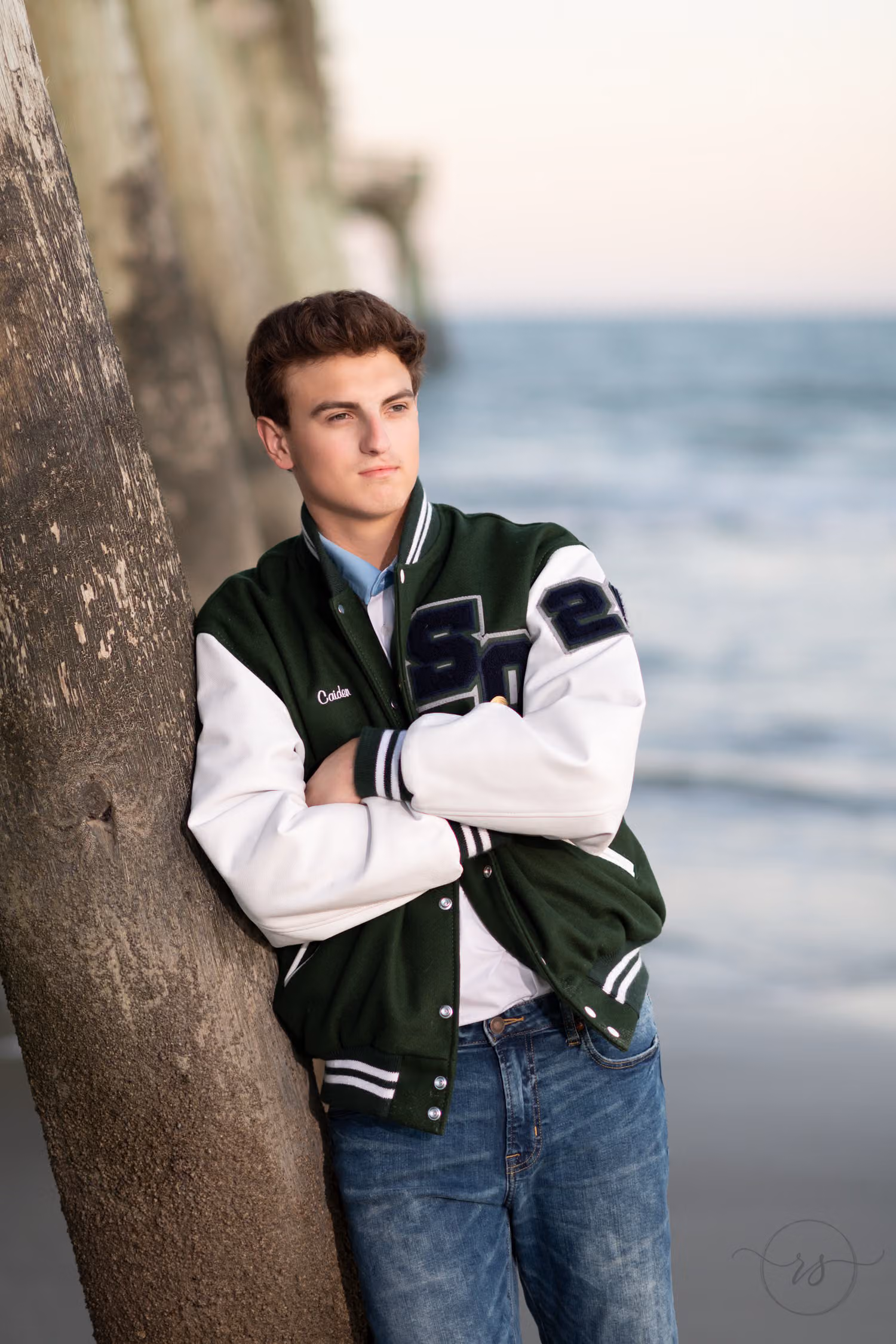 Senior with his arms crossed in a letterman jacket leaning on a pier piling at Myrtle Beach State Park