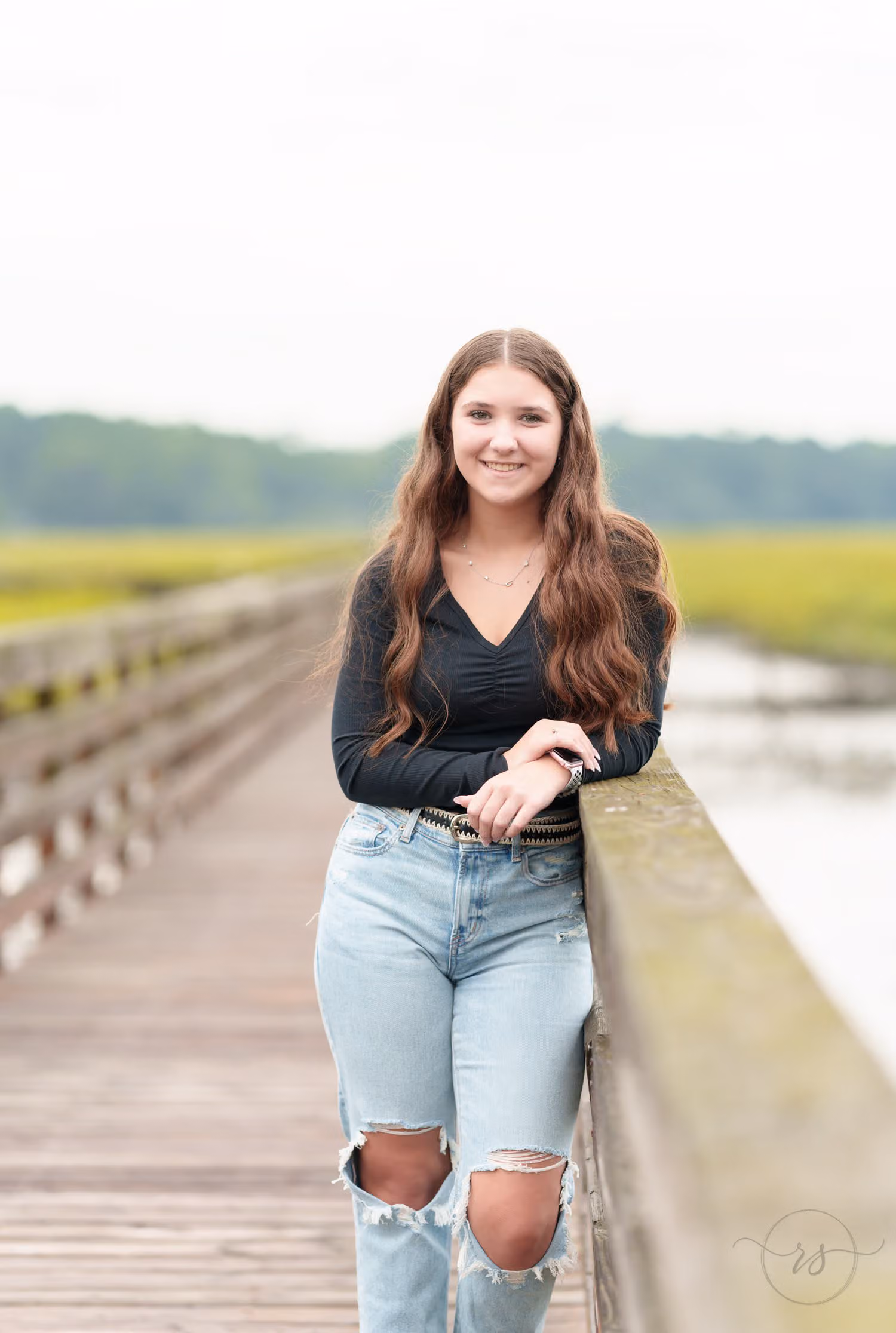 Senior in a black top and ripped jeans leaning on the boardwalk railing over the marsh at Huntington Beach State Park