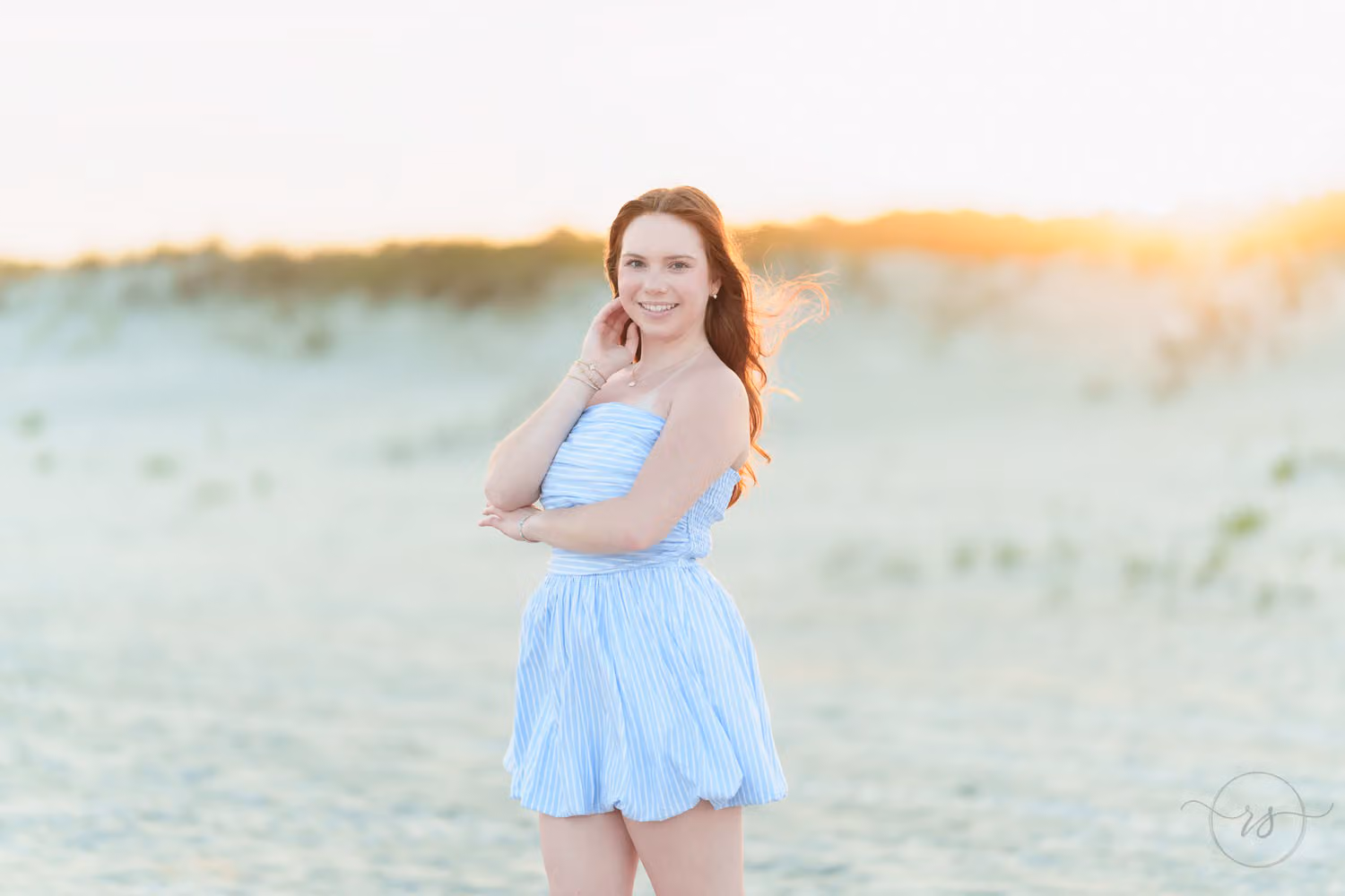 Senior girl in a light blue dress smiling by the dunes as the sun sets behind her at Huntington Beach State Park