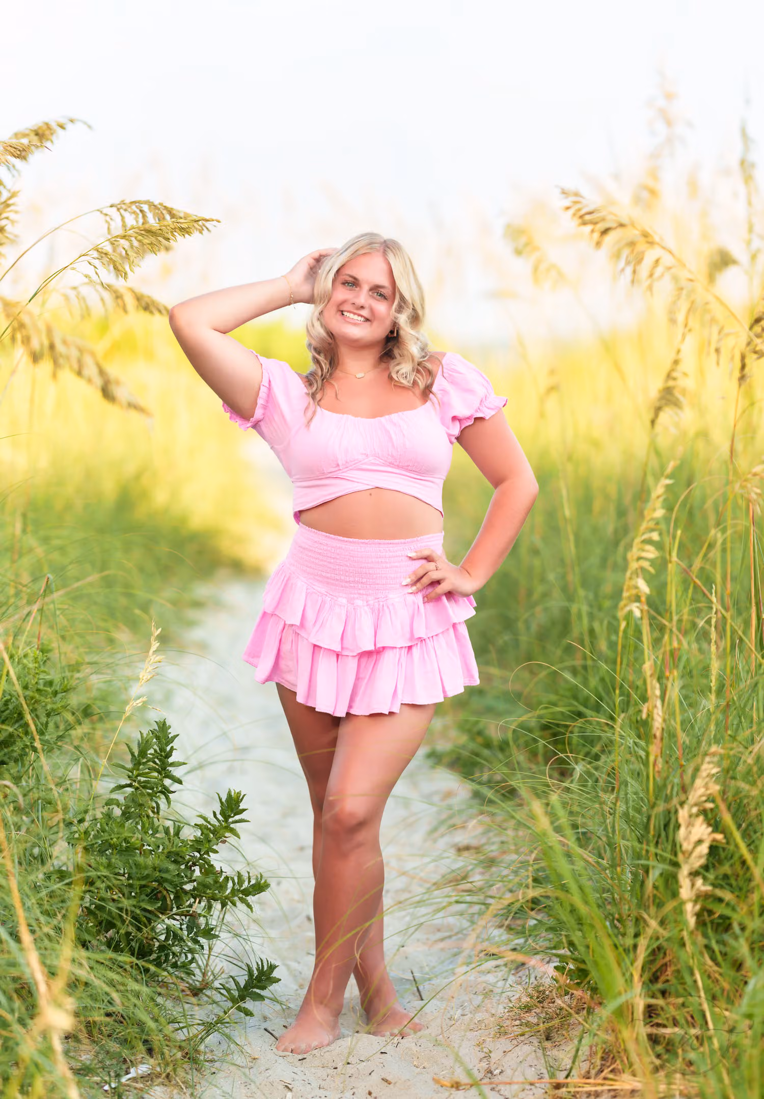 Smiling senior girl in a pink ruffled outfit standing on the sandy path between the sea oats in North Myrtle Beach