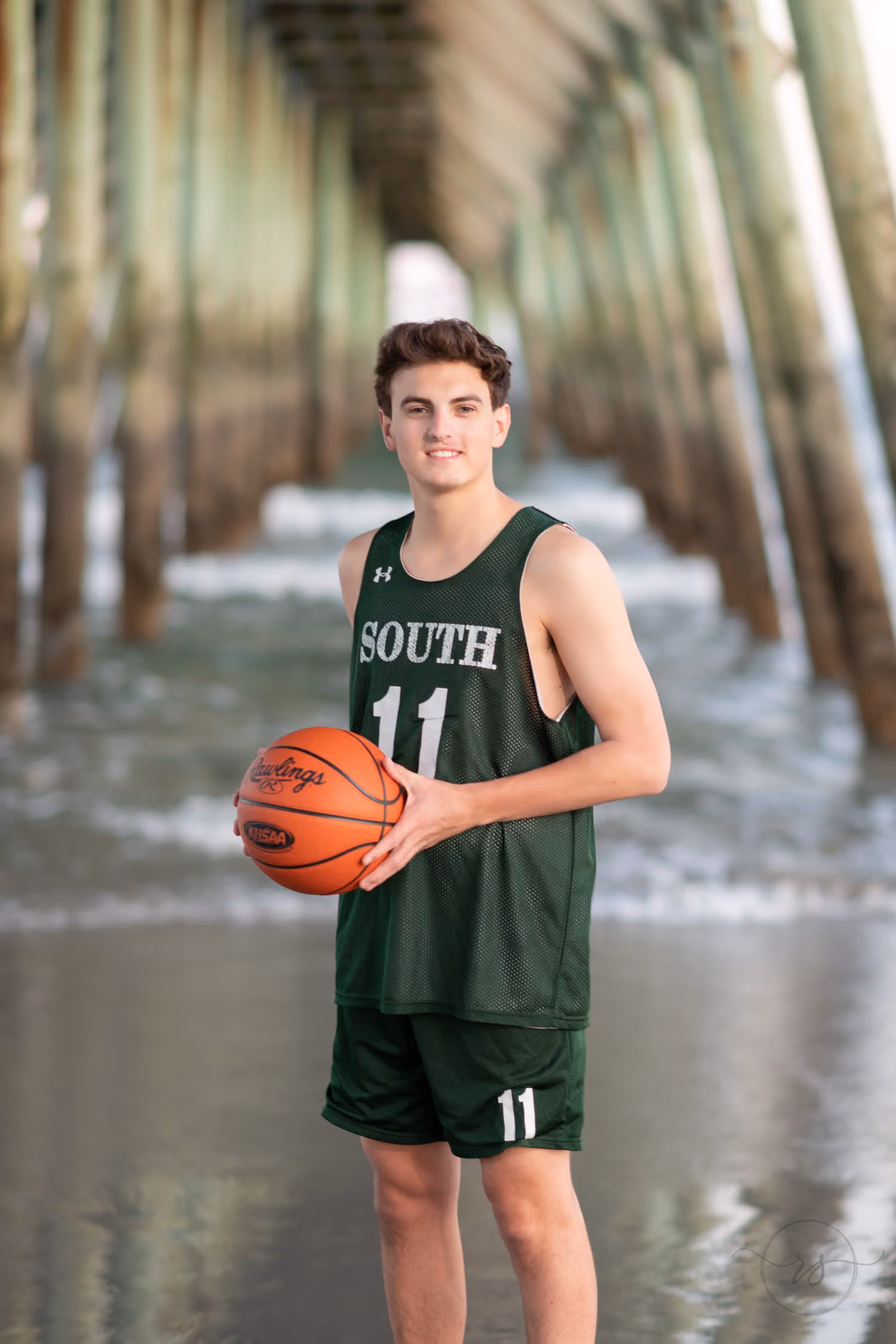 Senior in a green basketball uniform holding a ball in the shallow water under the pier at Myrtle Beach State Park