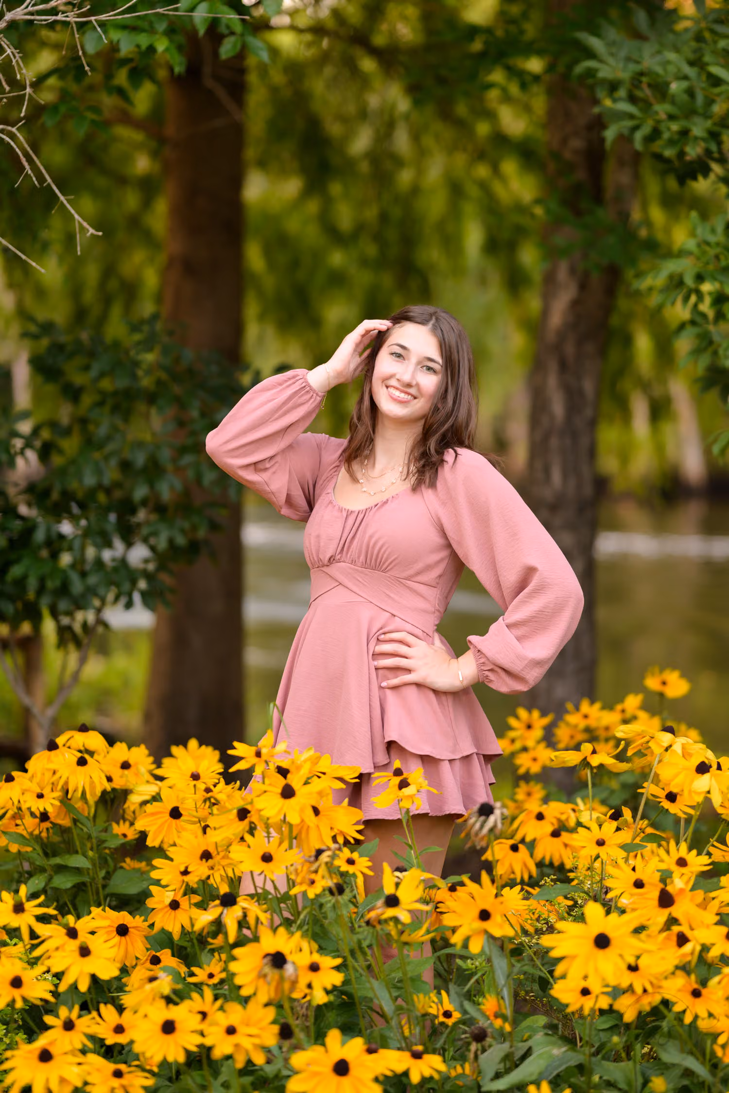 Senior girl in a pink dress standing behind a bed of yellow black-eyed Susans by the river at the Conway River Walk