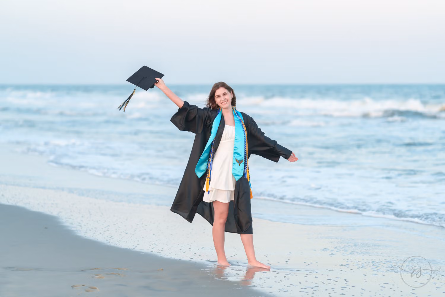 Senior girl in a black graduation gown happily waving her cap in the air by the ocean at Huntington Beach State Park